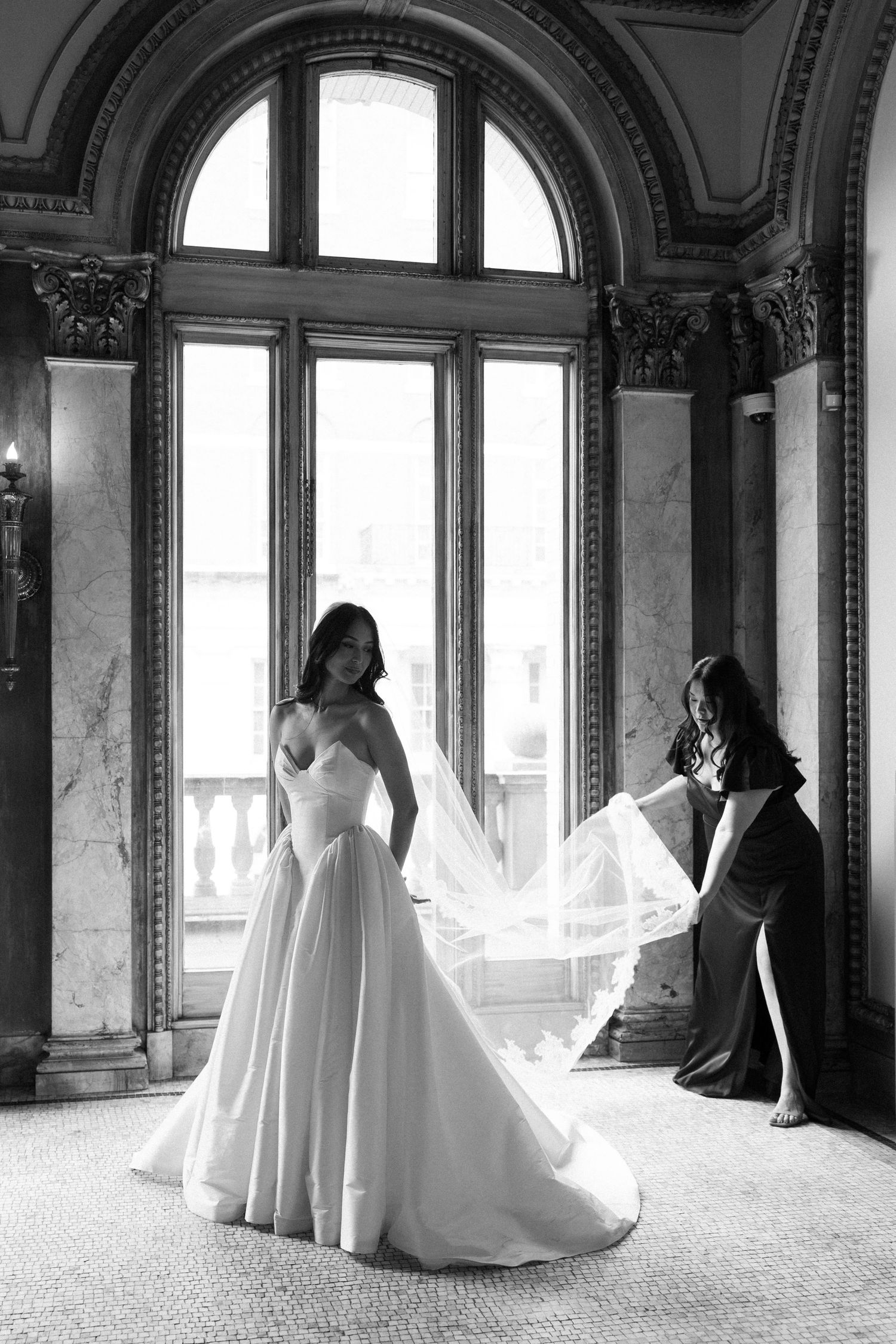 Black and white photo of a bride in a ballgown with veil being arranged by bridesmaid in an elegant venue.