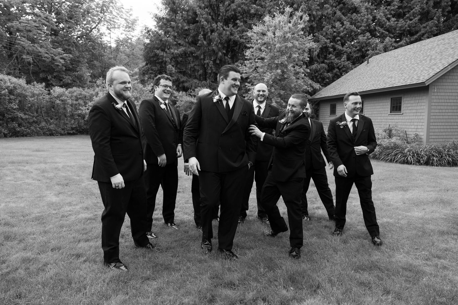 Black and white photo of a groom and groomsmen laughing together outdoors in formal suits at a wedding.