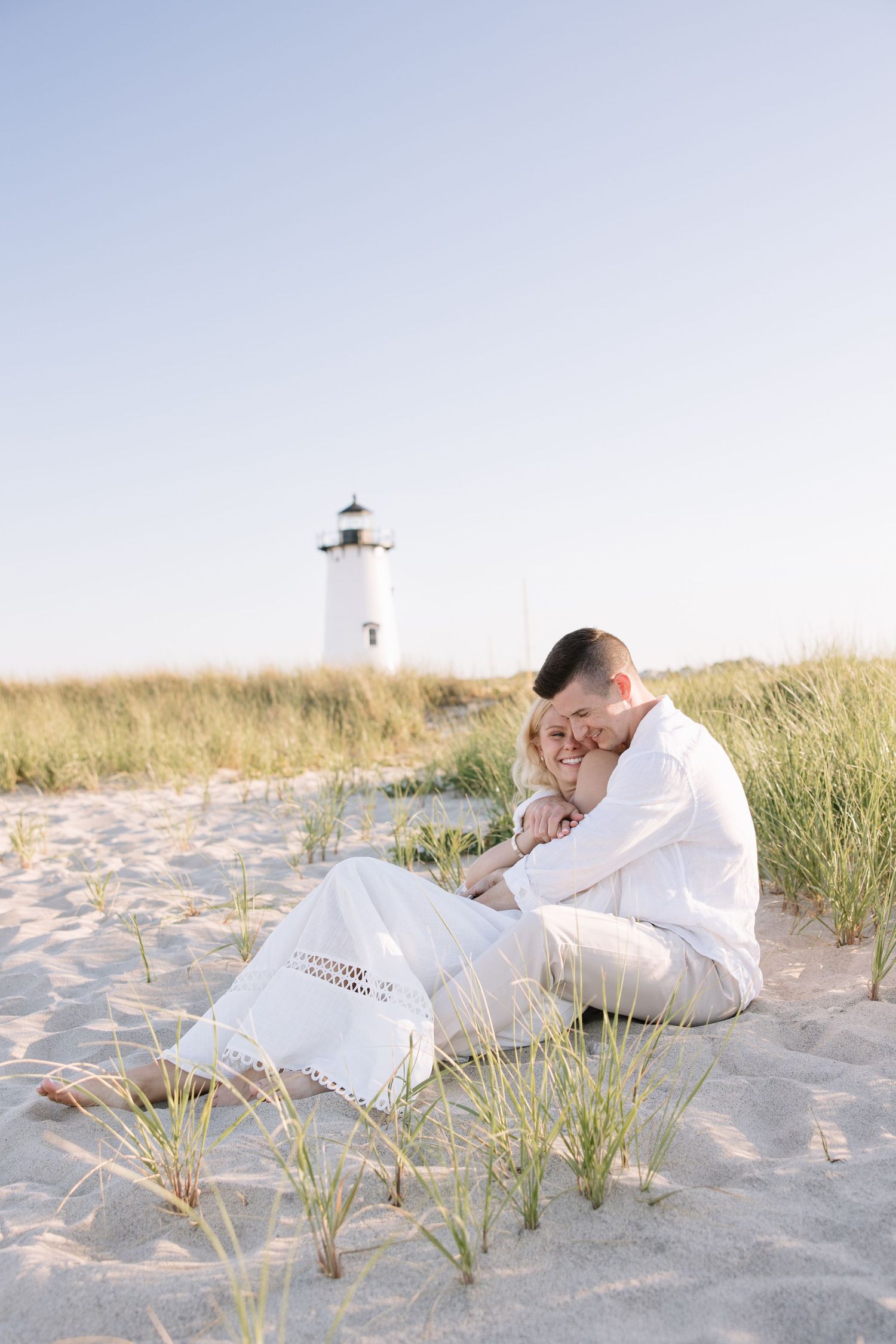 Couple in white clothing embracing on sandy beach dunes with a lighthouse in the background on a clear day.