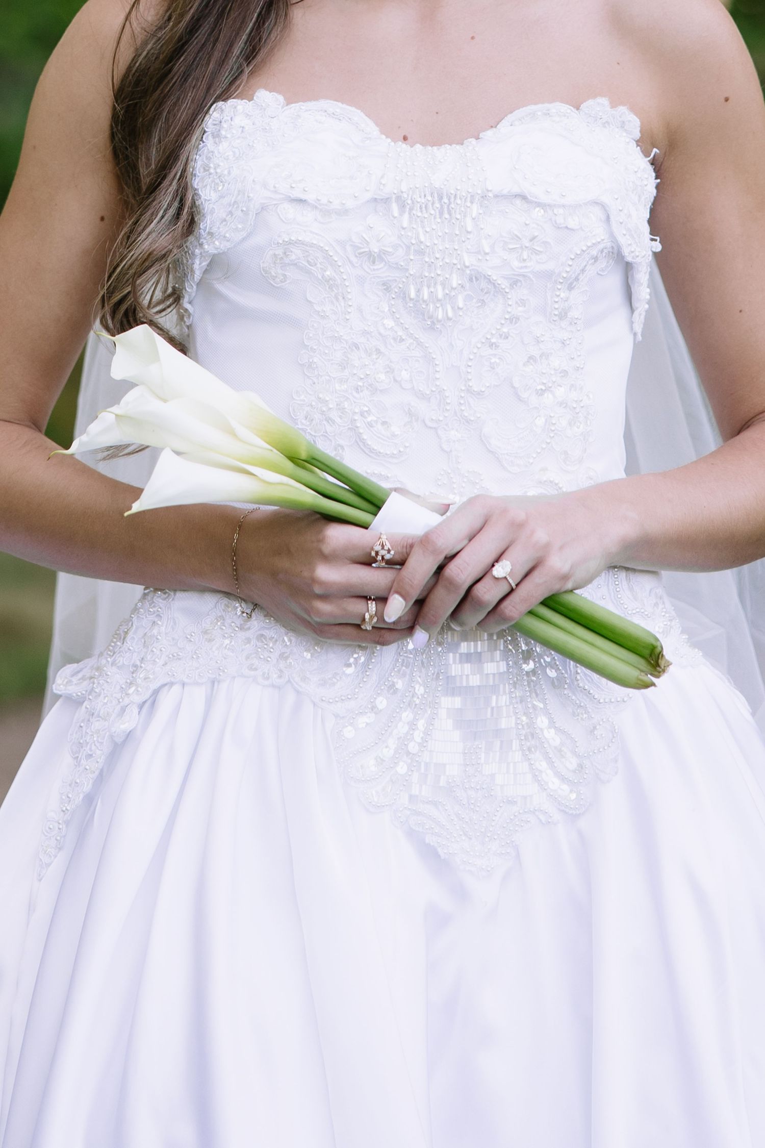 Bride in strapless white lace wedding gown holding elegant white calla lily bouquet tied with ribbon.