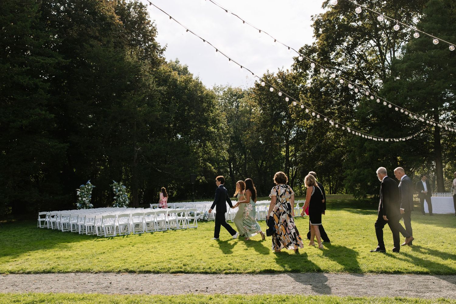Guests arrive at an outdoor wedding ceremony with white chairs and string lights in a lush green forest setting.