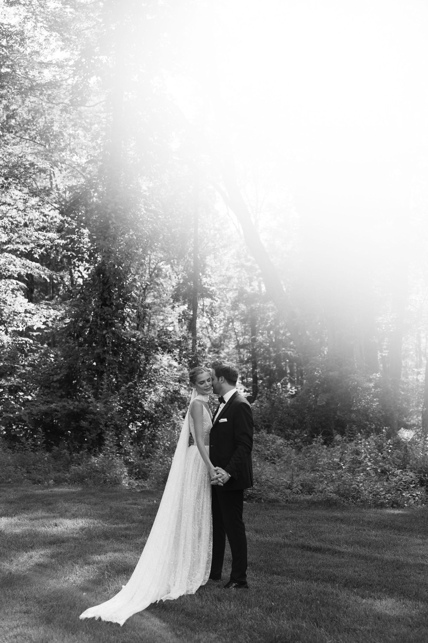 Black and white photo of bride and groom standing in sunlit forest clearing, bride in flowing gown with long veil.