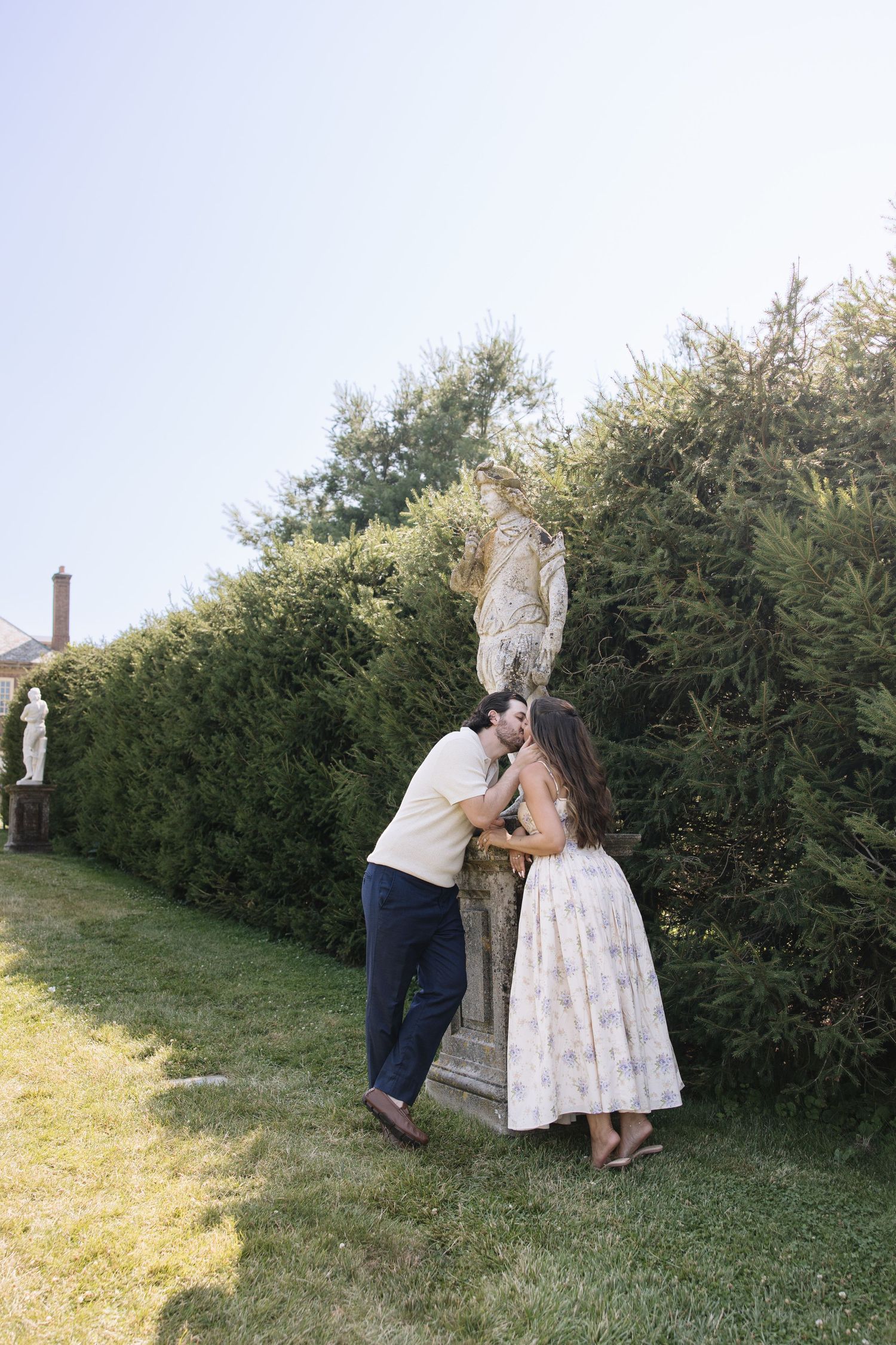 Couple sharing a romantic kiss beside a stone statue in a lush garden setting.