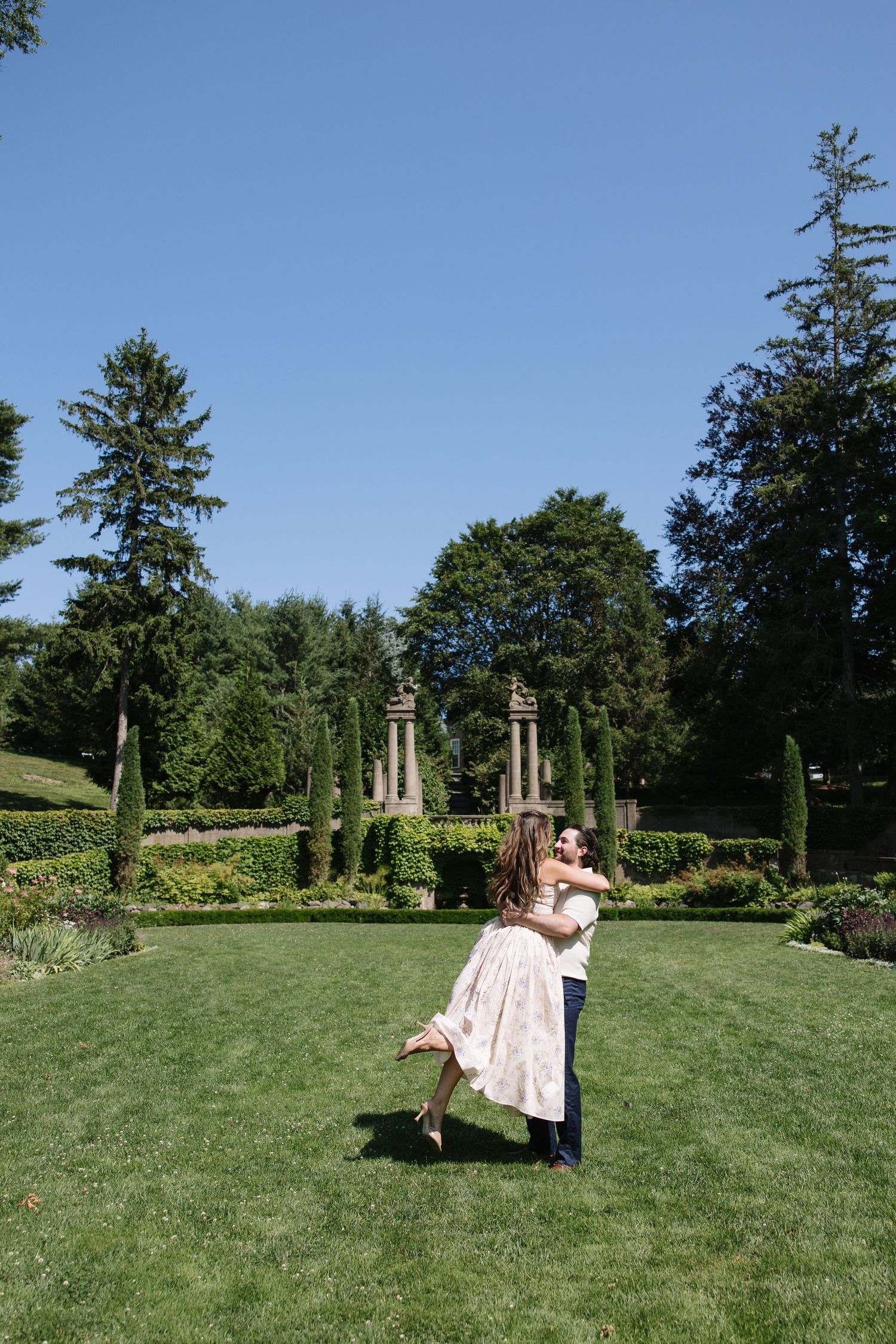 Couple dancing and embracing on lush green lawn in elegant garden with stone pillars and tall trees under clear blue sky.