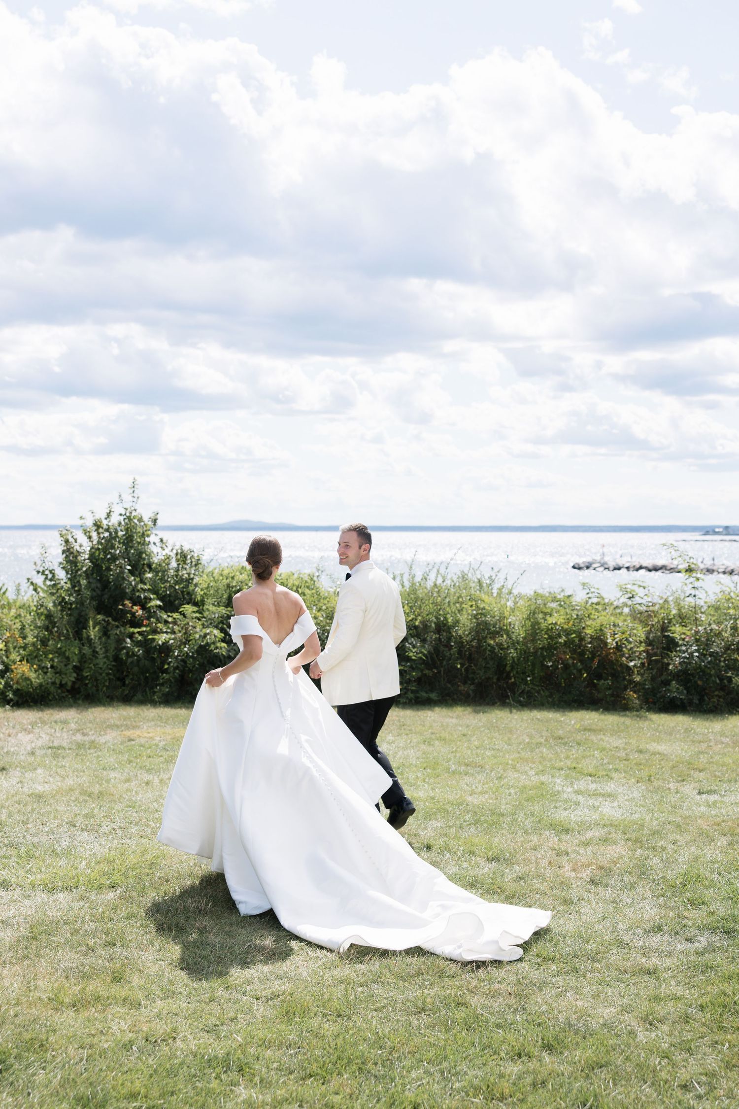 Bride and groom walking hand in hand on green lawn overlooking the ocean on their wedding day.