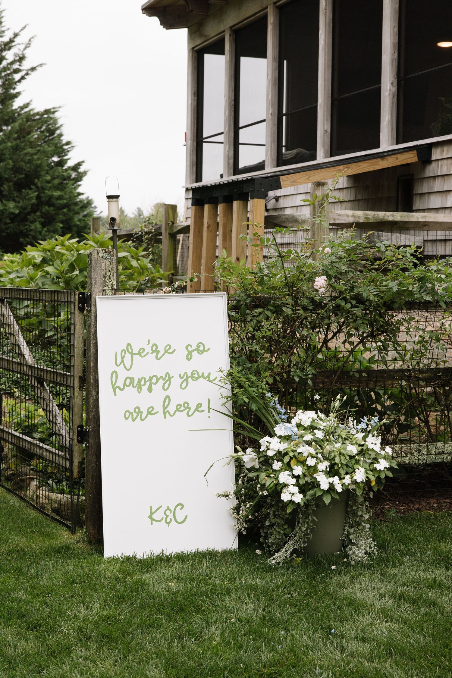 Outdoor wedding welcome sign reading We're so happy you are here with initials K&C, beside a floral arrangement on green lawn.
