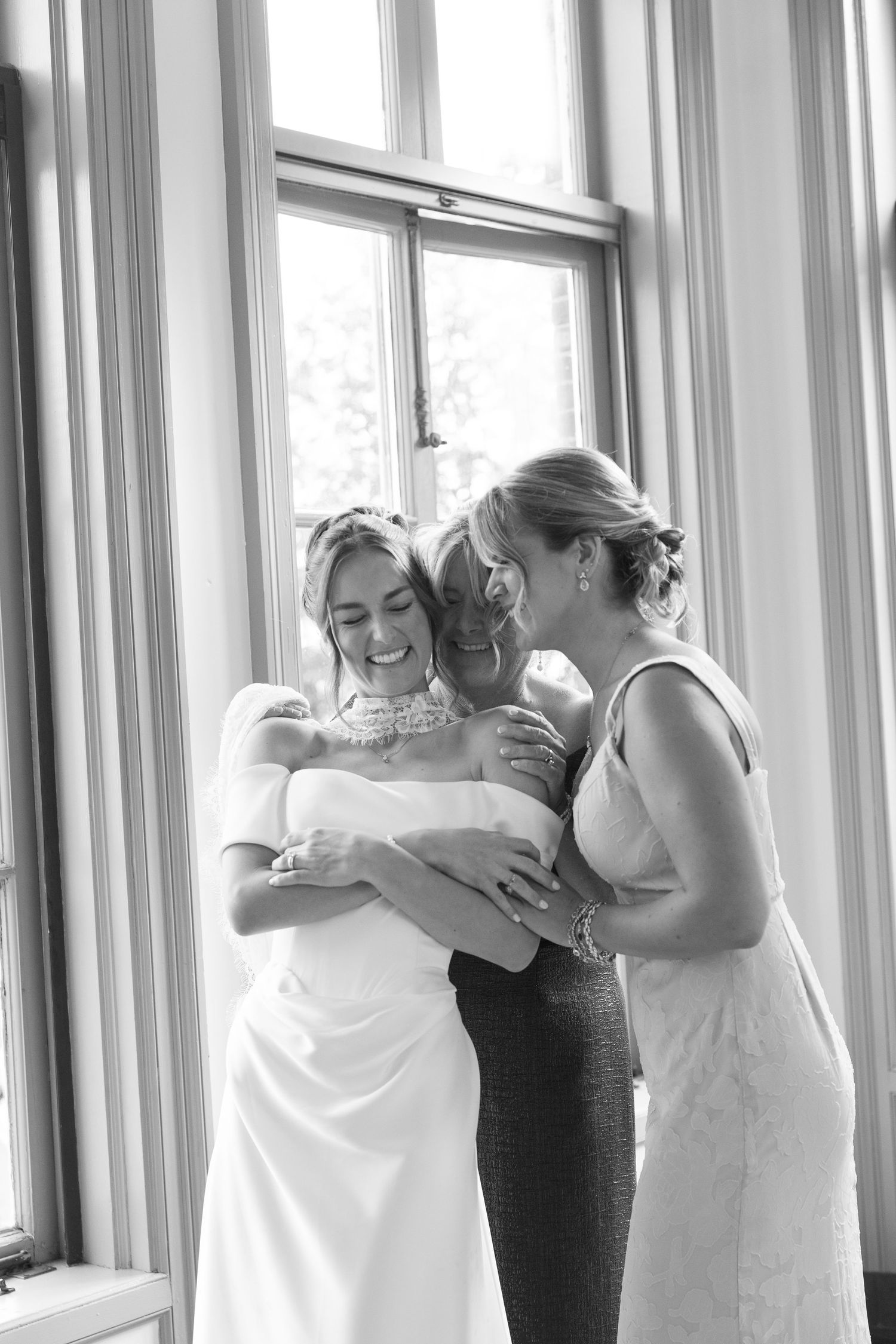 Black and white photo of bride sharing emotional embrace with two women near bright windows before wedding.