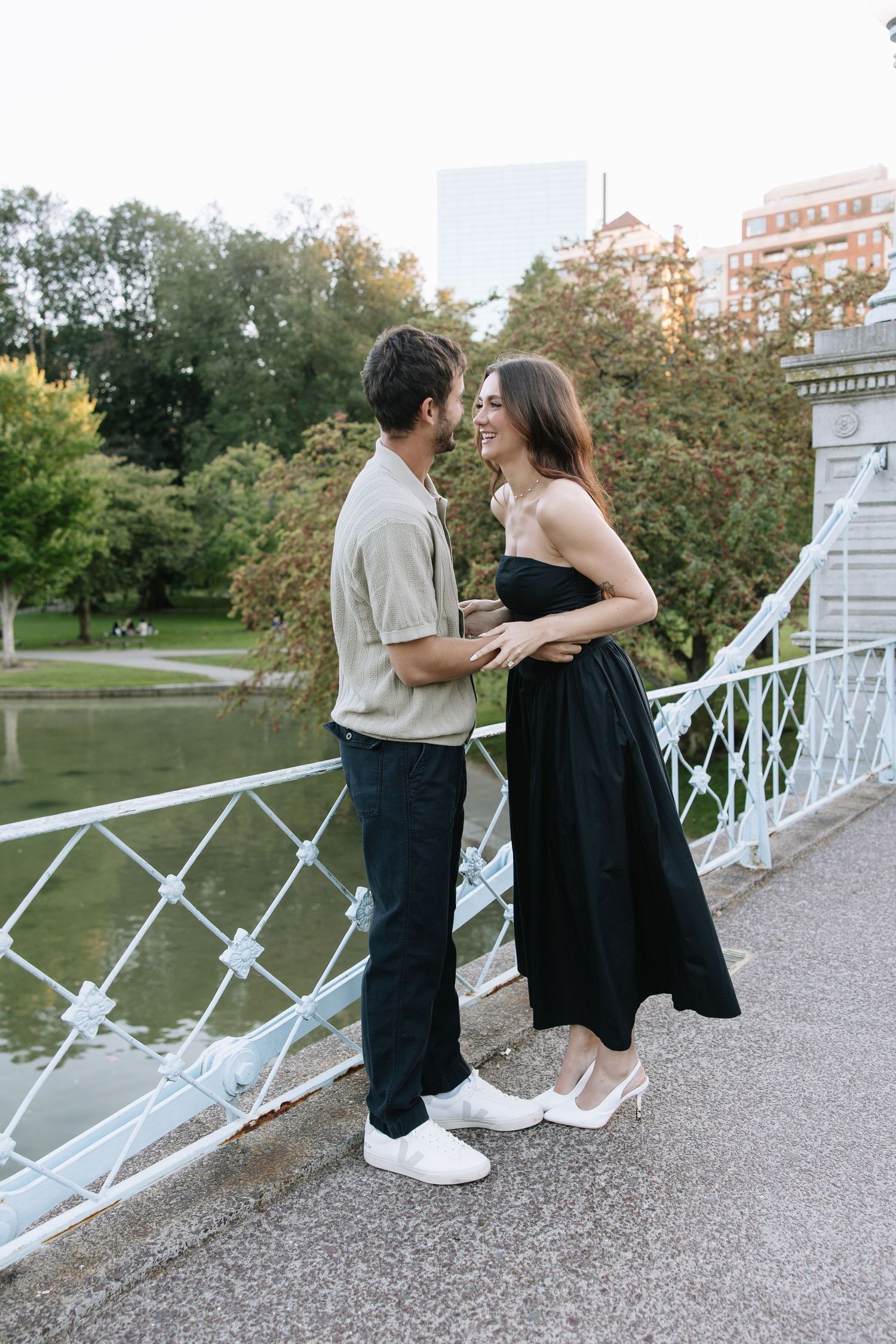 Two people share a romantic moment on a white bridge overlooking a park.