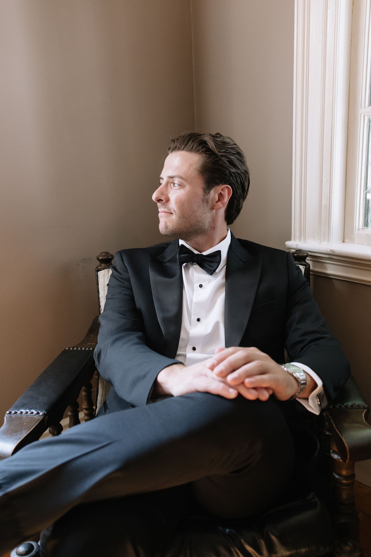 Groom in classic tuxedo relaxing in chair during wedding preparations.