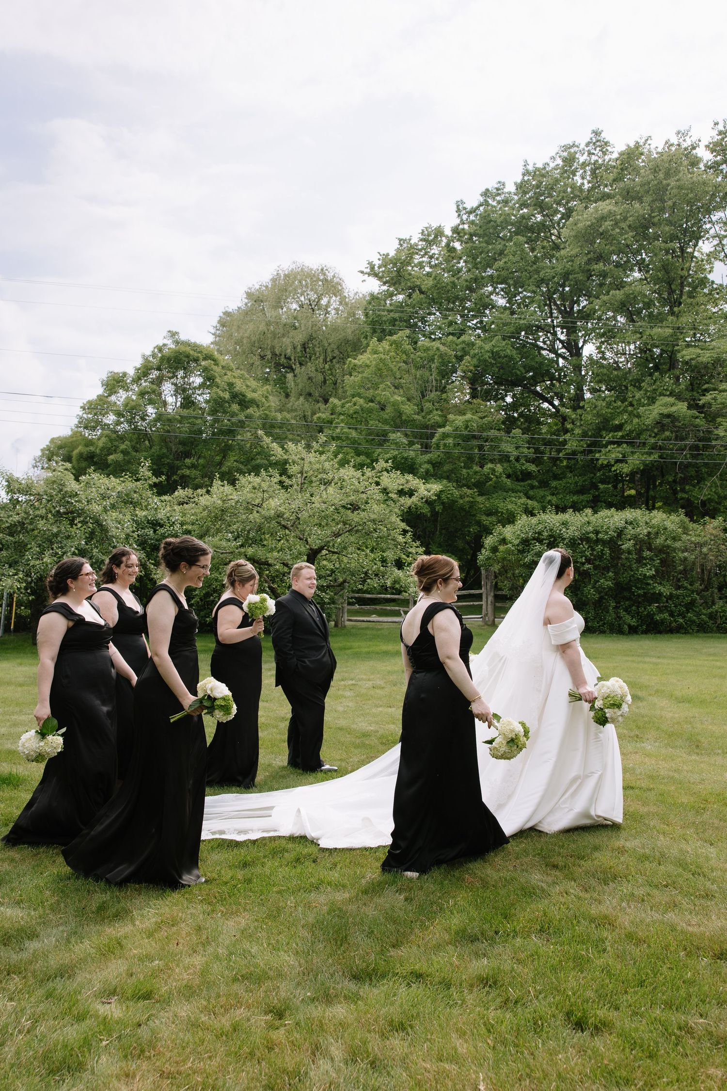 Bride in white gown with long veil walks with bridesmaids in black dresses across a green lawn.