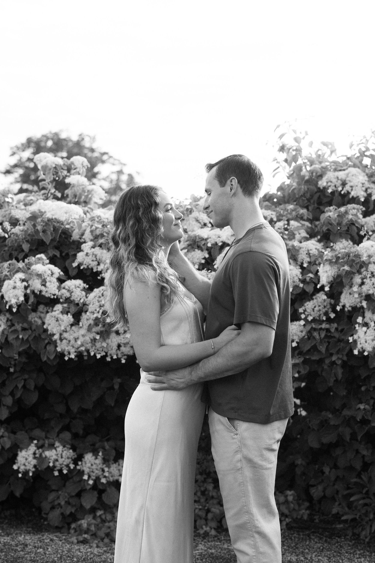 Black and white photo of a romantic couple embracing in front of blooming hydrangea bushes.