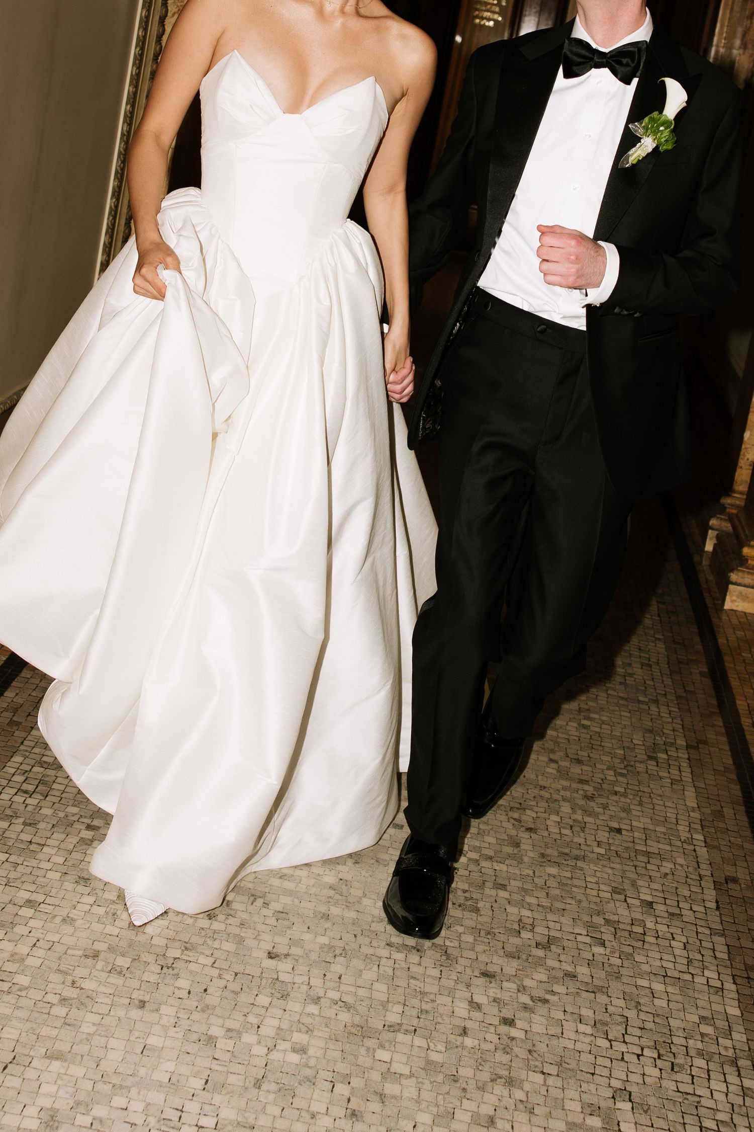 Bride in strapless white ballgown and groom in black tuxedo walking hand in hand at wedding.