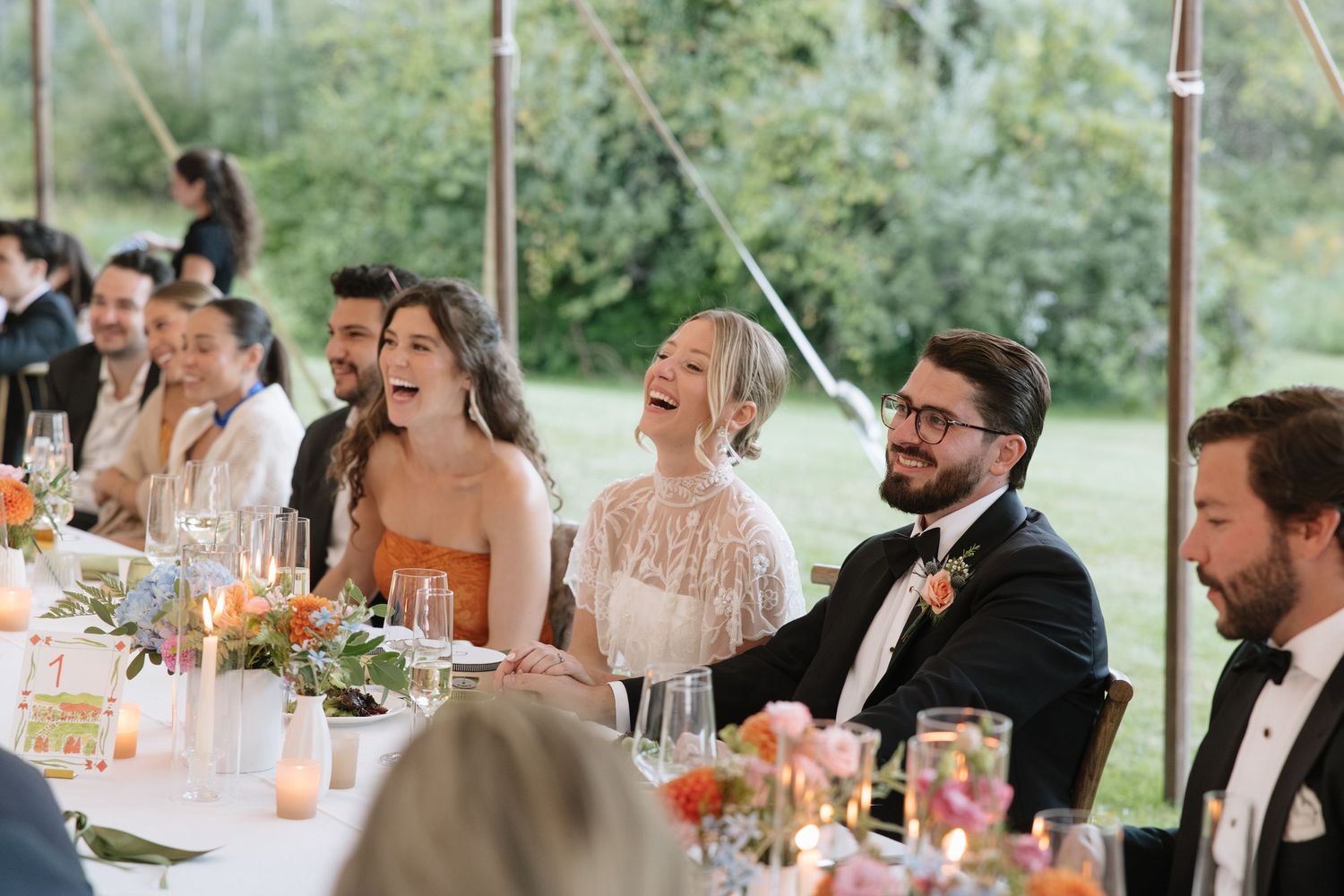 Wedding guests laughing joyfully at head table during reception speeches under an elegant open-air tent.