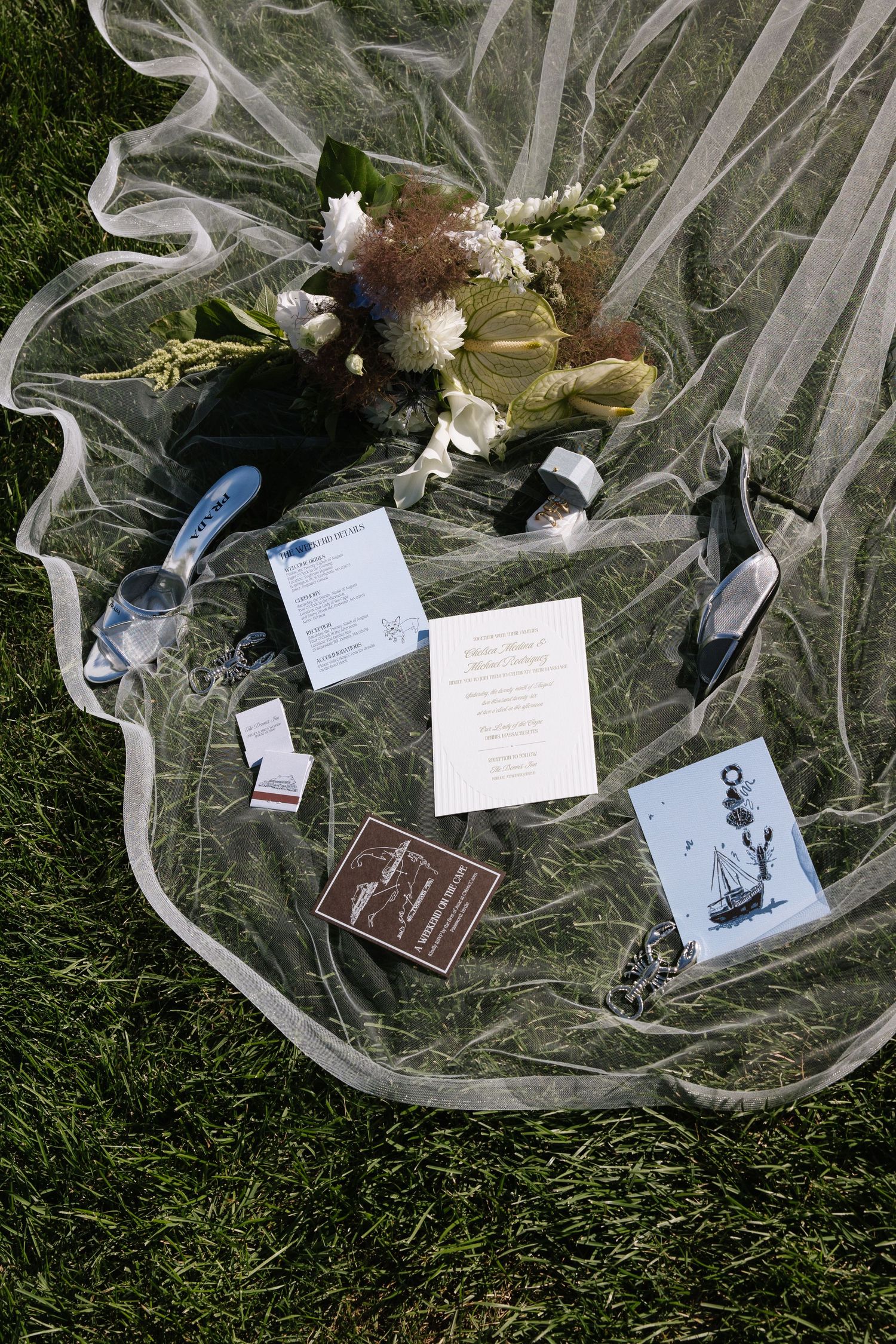 Wedding flat lay with veil, bouquet, invitation suite, shoes, and accessories arranged on green grass.