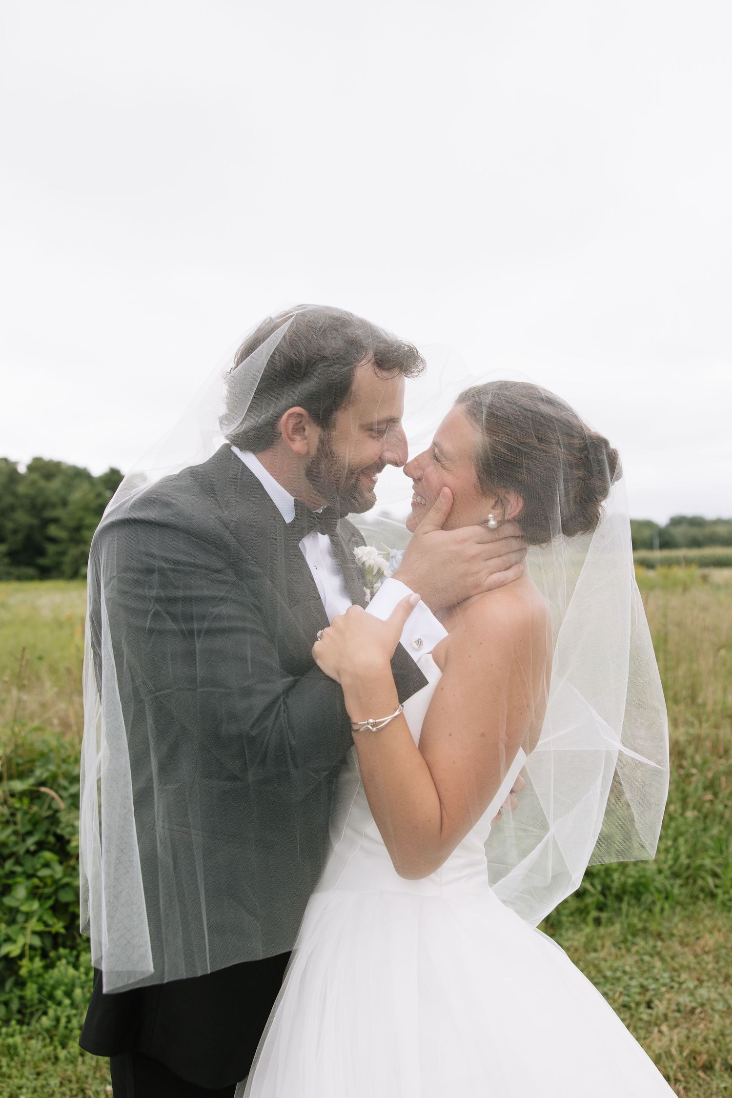 Bride and groom share intimate moment under wedding veil in a green field on their wedding day.