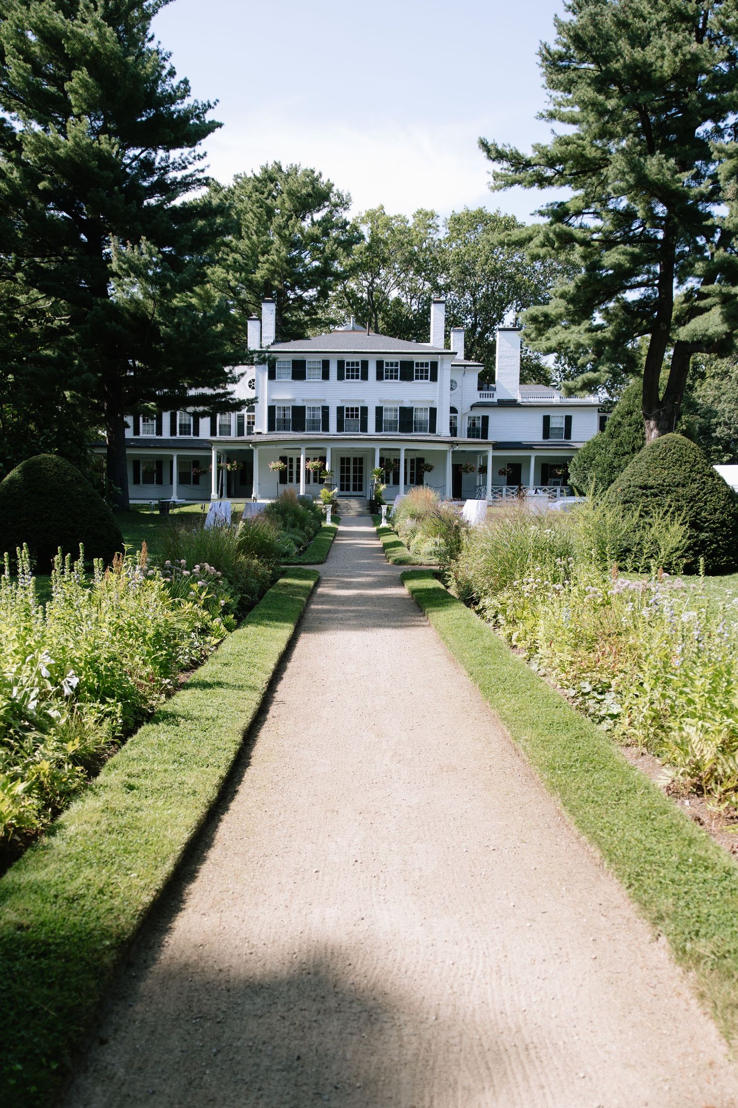 White colonial style mansion with manicured gardens and gravel pathway leading to entrance.