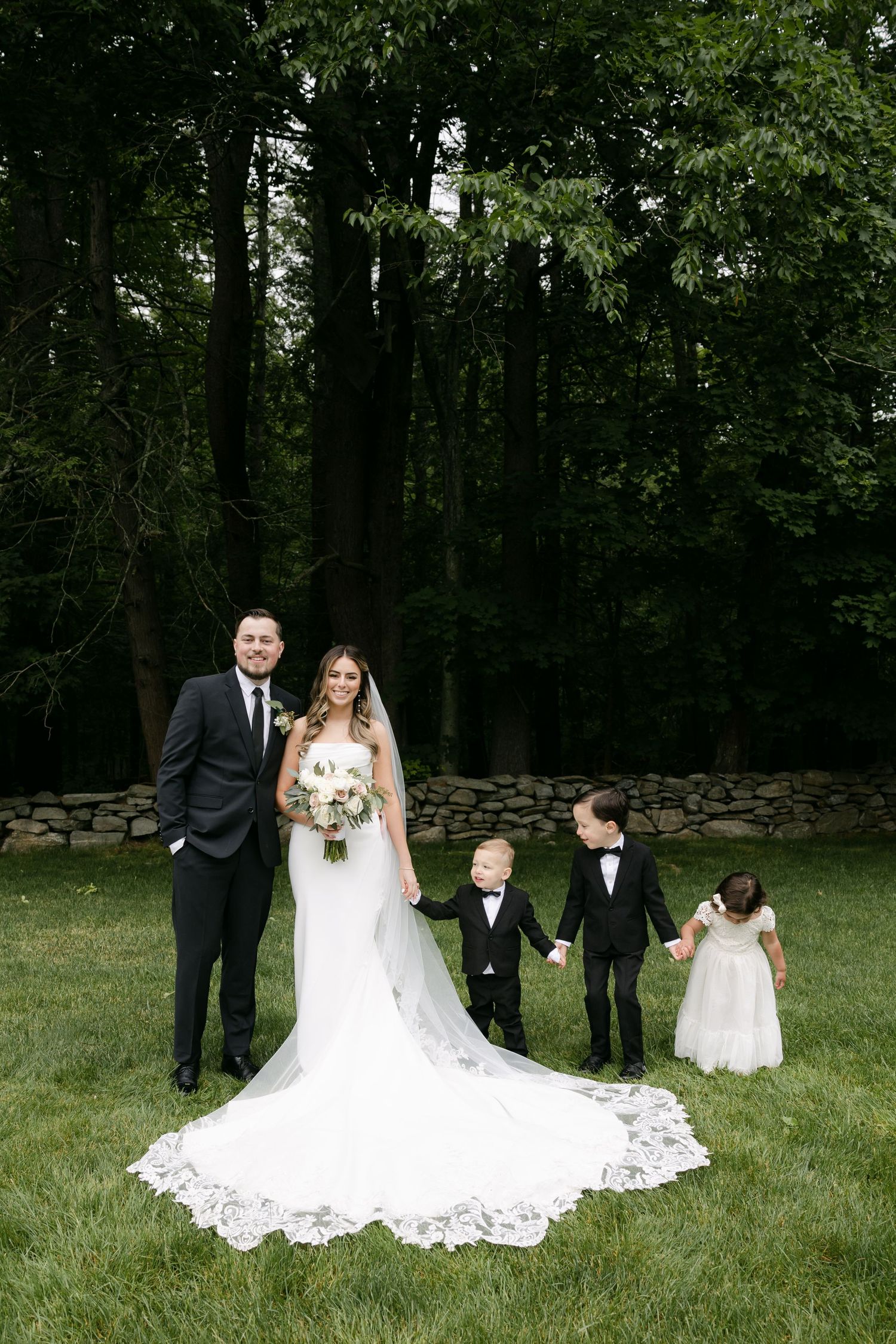 Outdoor wedding portrait with flower girls and ring bearers on grassy lawn.