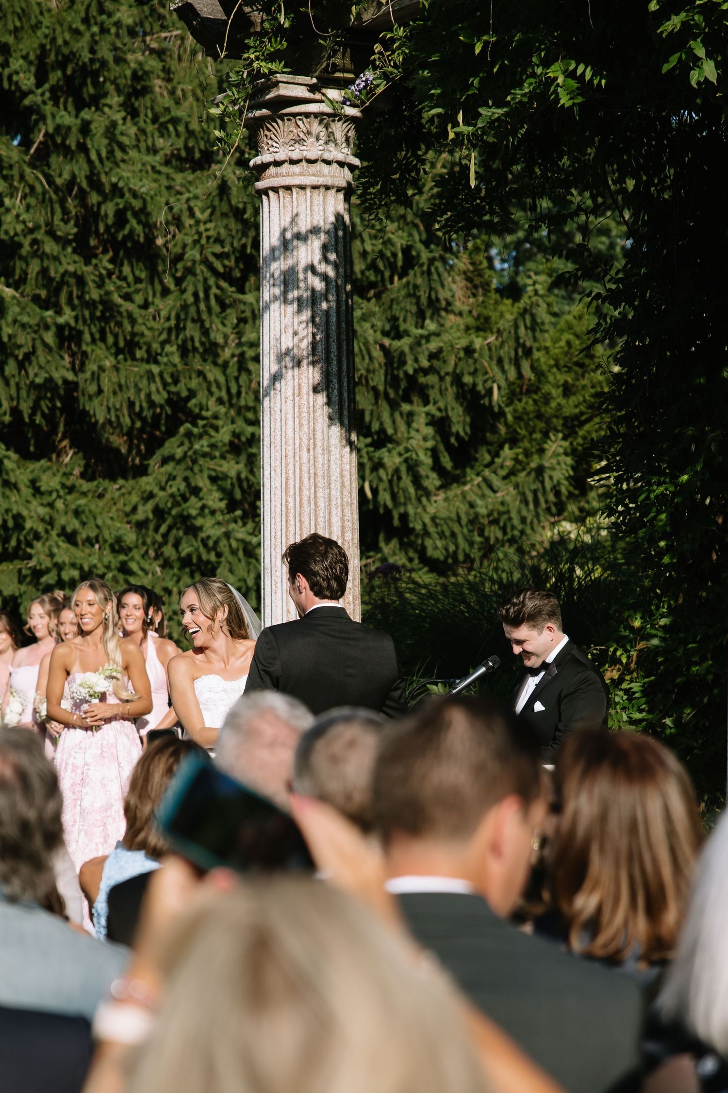 Wedding ceremony in progress with guests seated before classical columns in a garden setting.