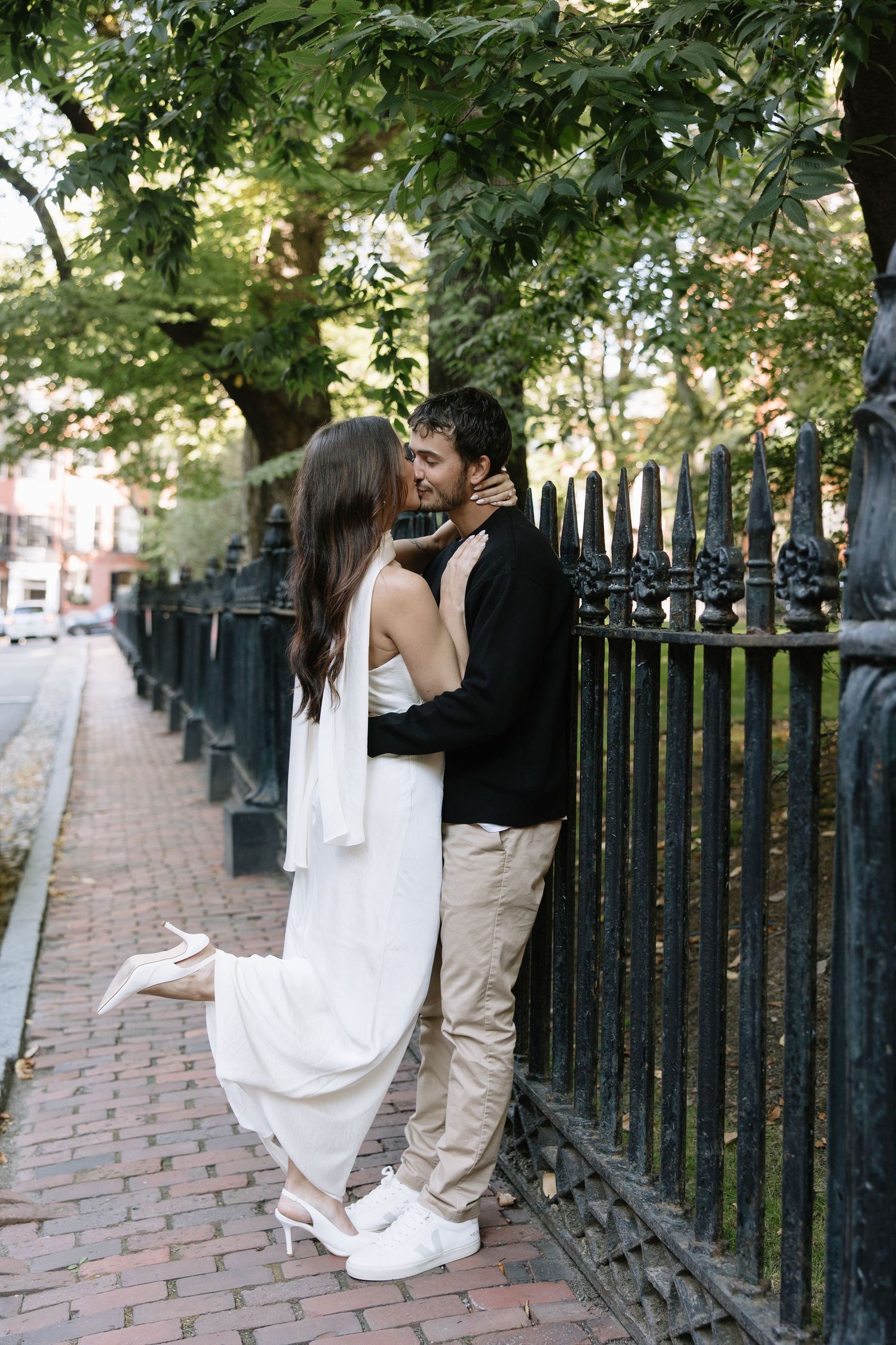 A couple shares a romantic moment against an iron fence on a brick-lined street.