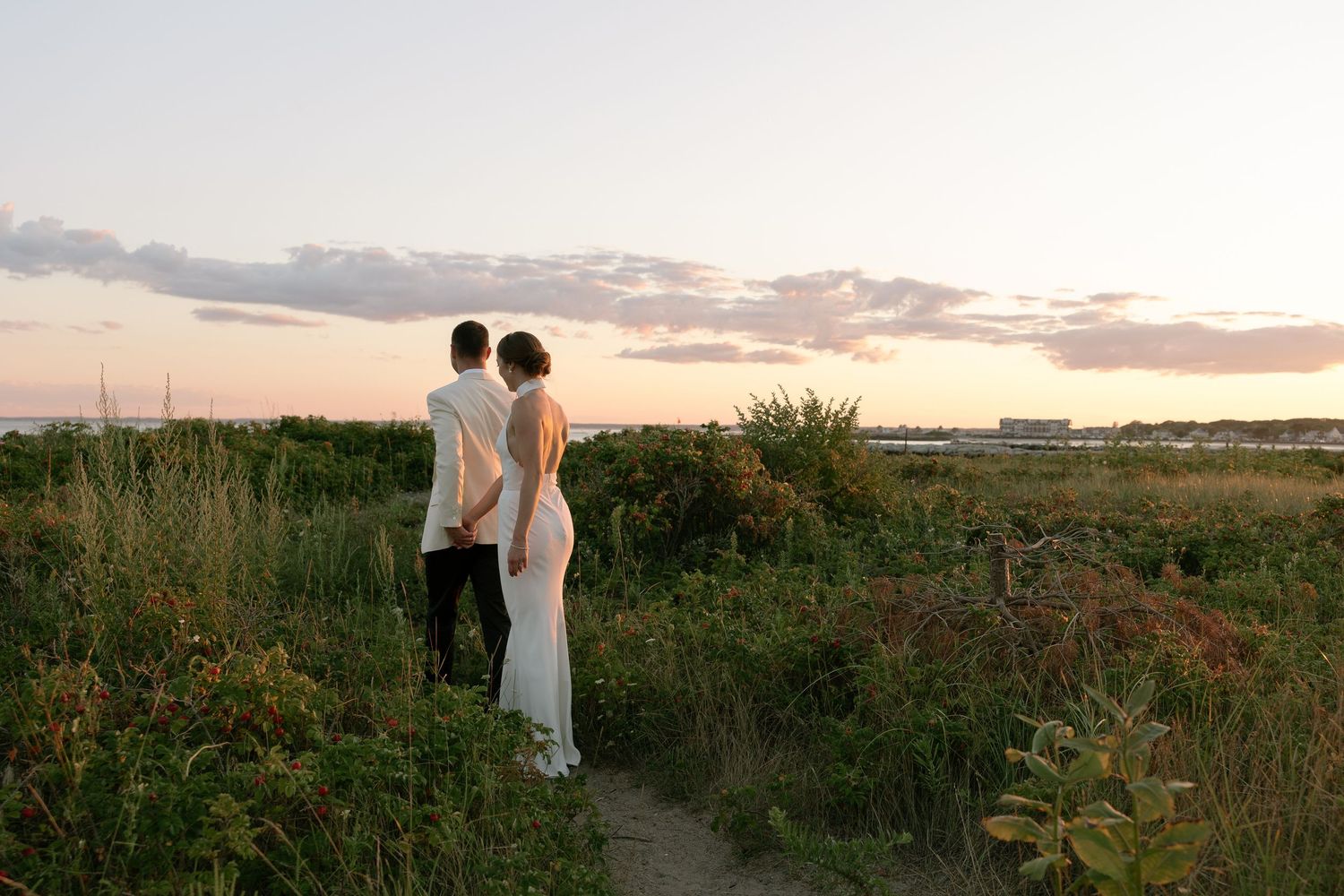 Bride in a white gown and groom in a light jacket stand holding hands on a coastal path through grass and shrubs at sunset.