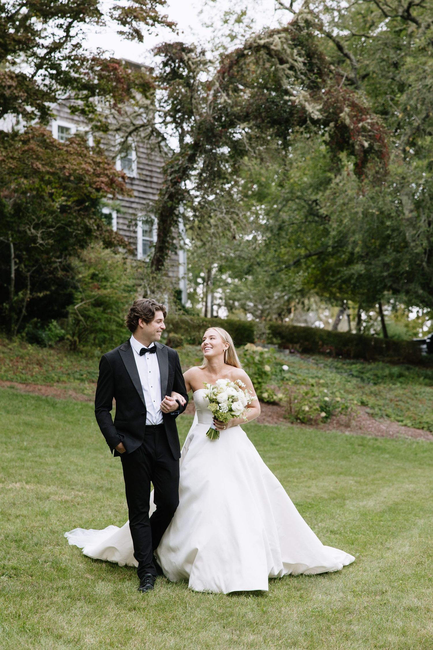 Bride and groom walking together on a green lawn in front of a charming estate surrounded by lush trees.