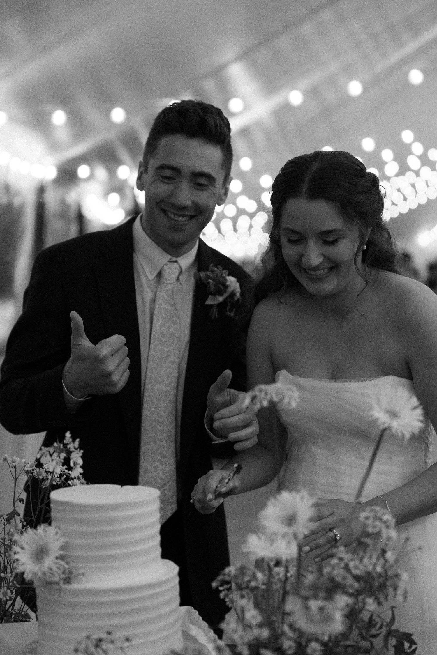 Black and white photo of smiling bride and groom cutting their tiered wedding cake at elegant reception.