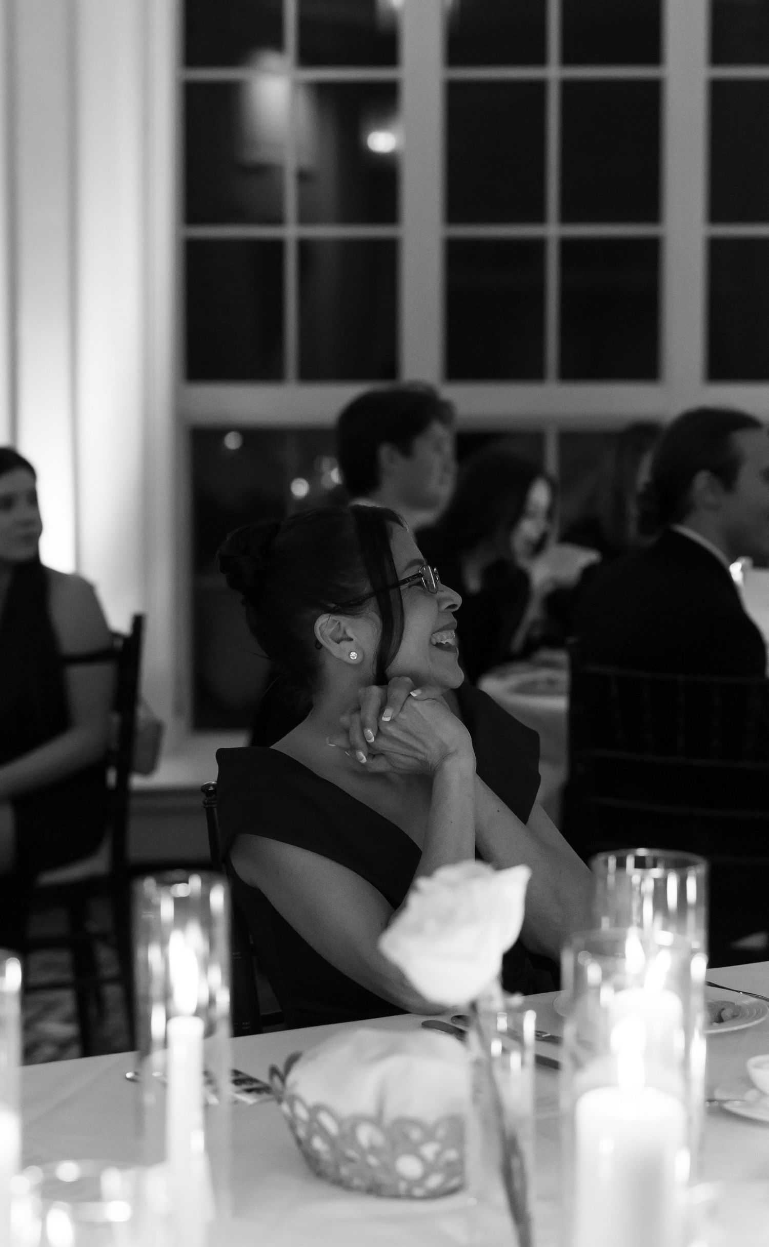 Black and white photo of a woman laughing at a candlelit formal dinner event with guests in the background.