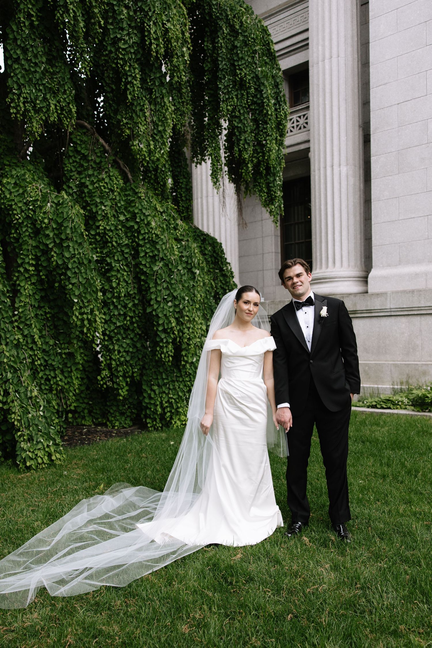 Bride and groom pose on green lawn beside ivy-covered building on wedding day.