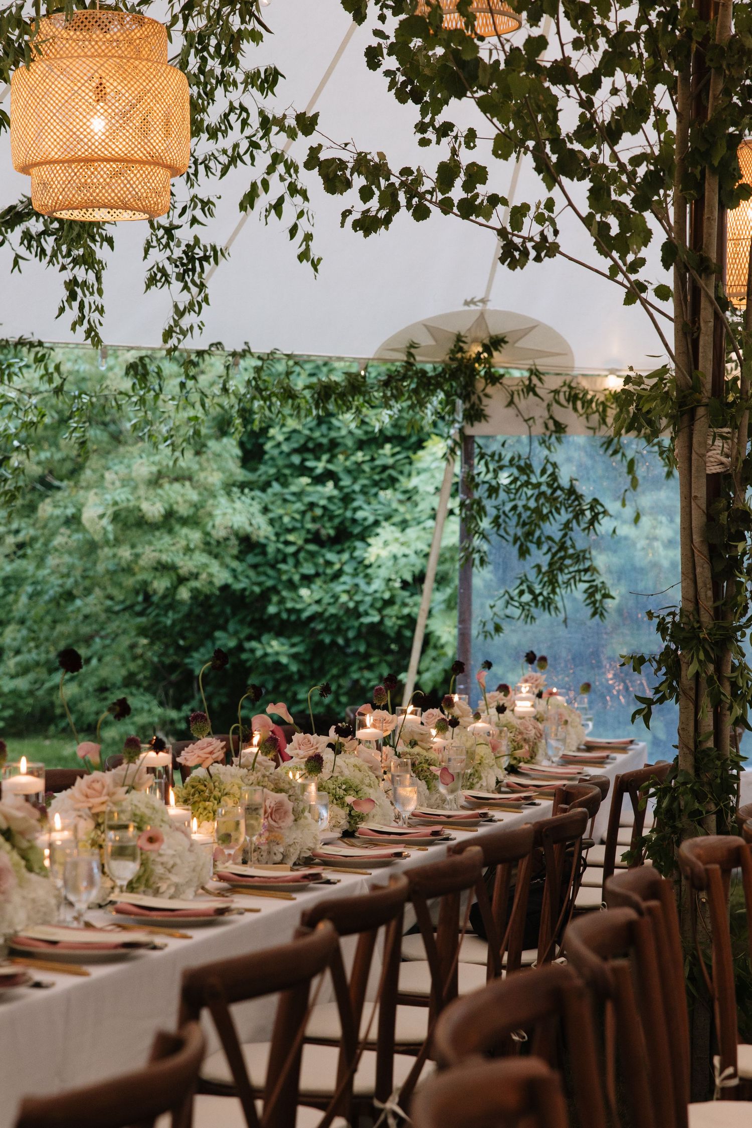 Elegant outdoor wedding reception with long dining tables, floral centerpieces, candles, wooden chairs, and lush greenery under a tent.