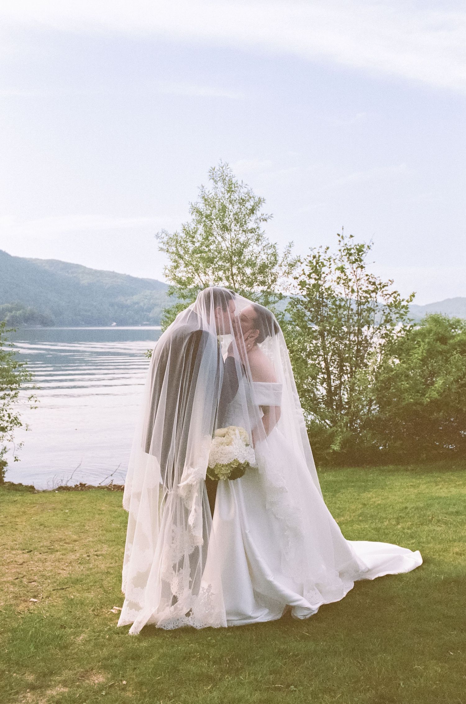Bride and groom sharing a kiss under a flowing veil by a lakeside, surrounded by lush greenery and mountains.