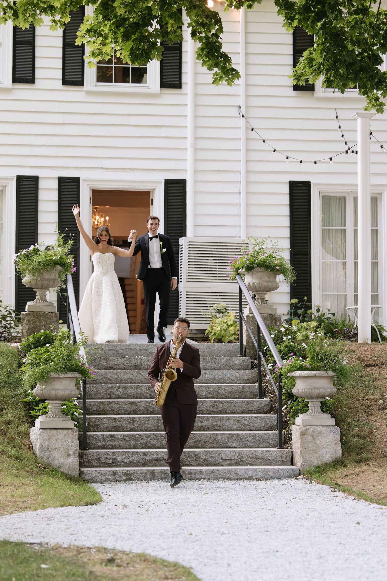 Bride and groom celebrate on white colonial home steps as saxophonist leads their wedding reception grand exit.