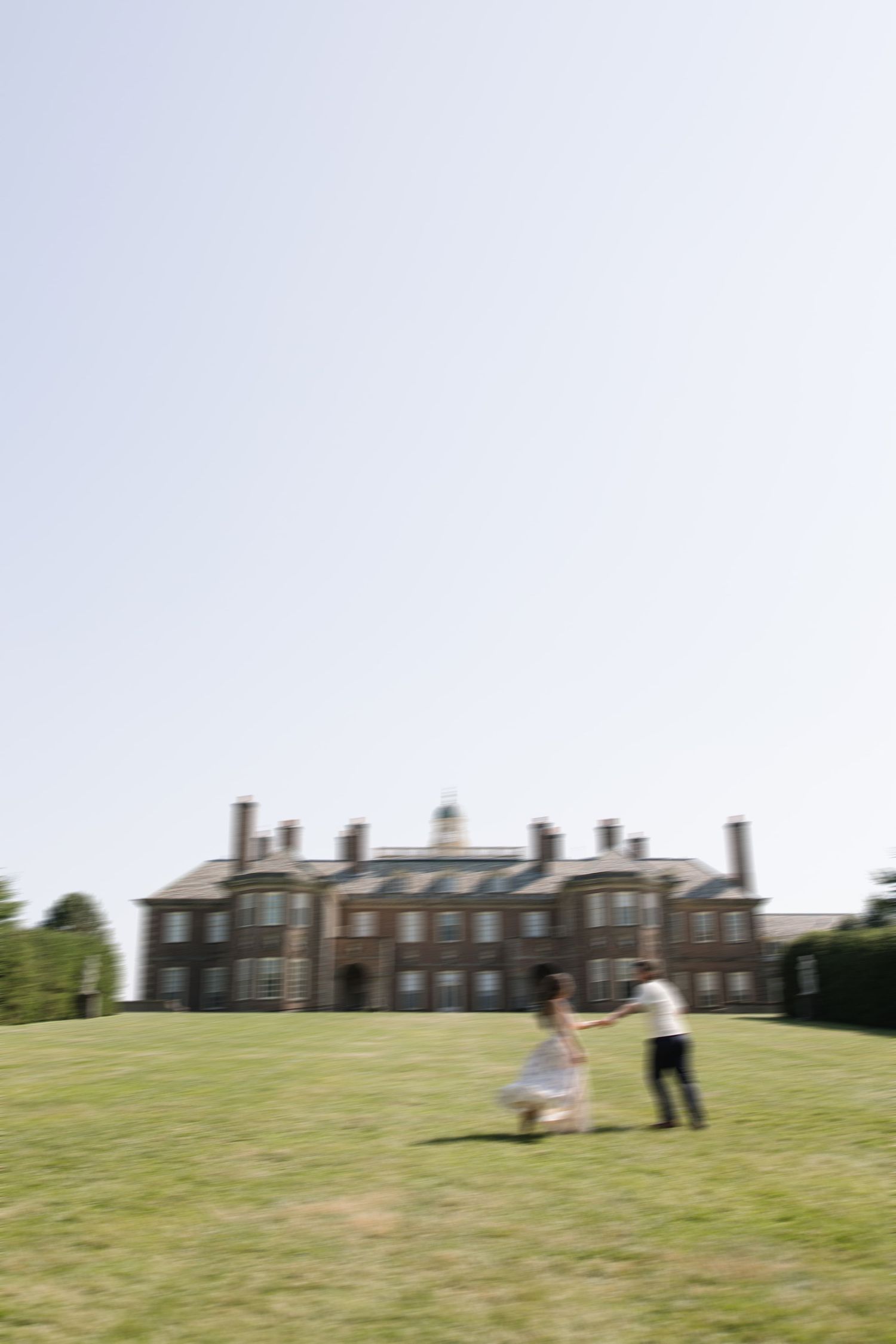 Couple holding hands and dancing on green lawn in front of a grand brick manor house on a sunny day.