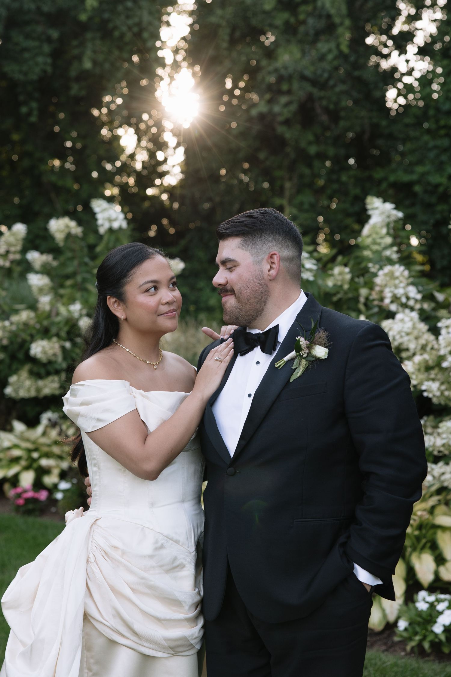 Bride in off-shoulder gown adjusts groom's bow tie in garden at sunset.