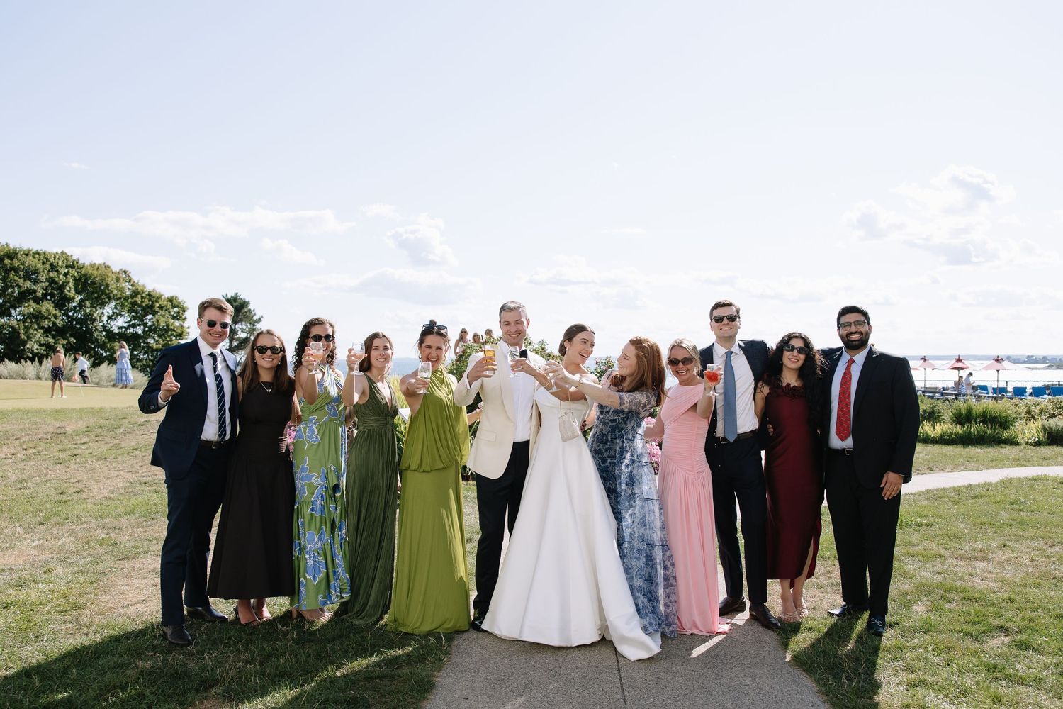 Wedding party celebrating outdoors, bride in white gown with guests in formal attire raising glasses on sunny day.