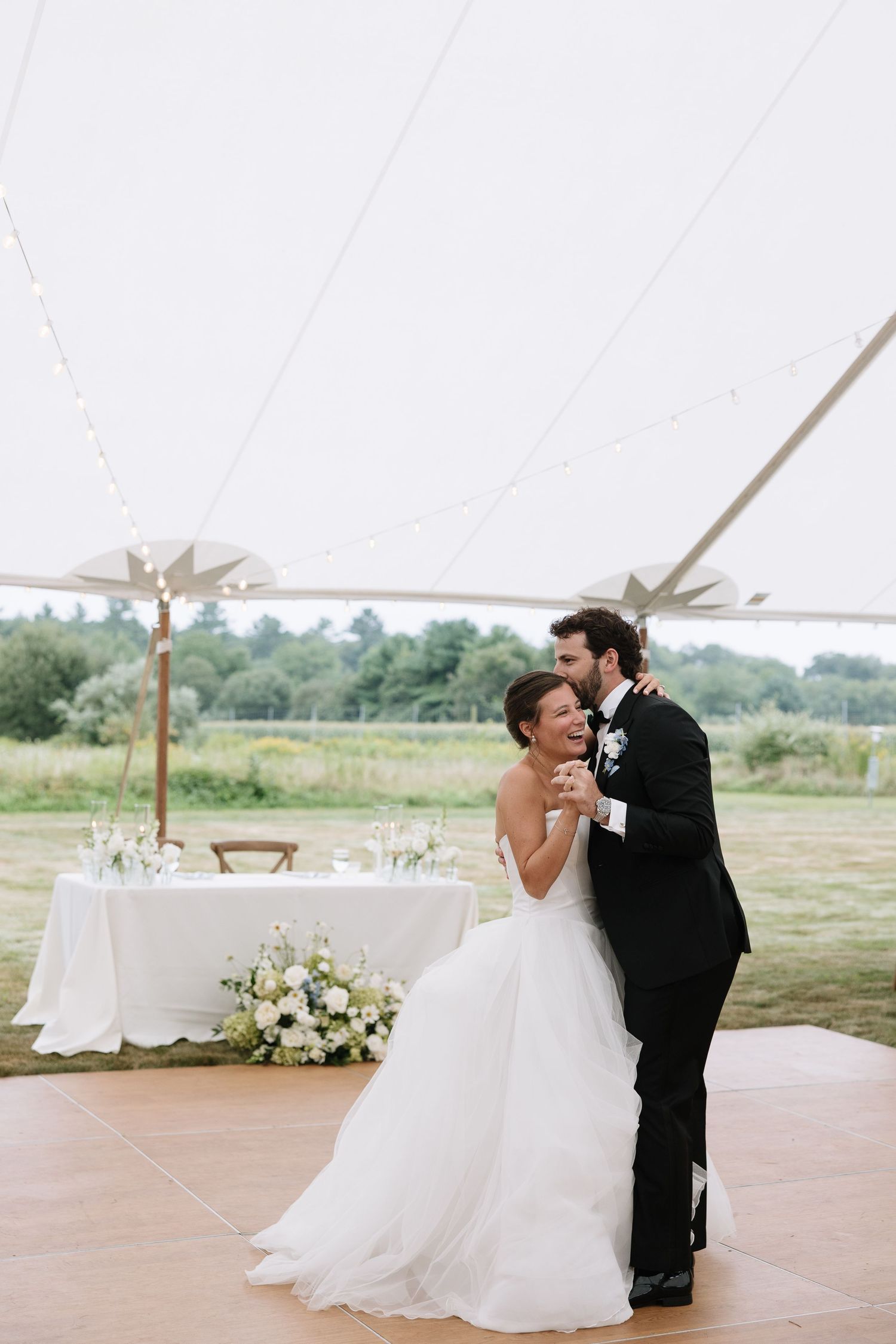 Bride and groom share first dance under elegant outdoor wedding tent surrounded by lush greenery.