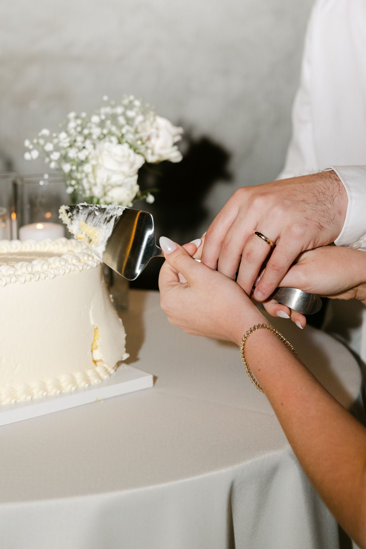 Hands carefully cutting into an elegant white wedding cake decorated with flowers.