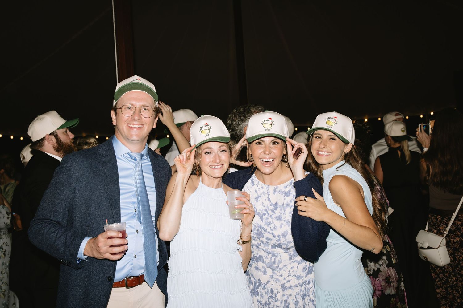 Group of guests wearing matching white baseball caps pose together at outdoor event.