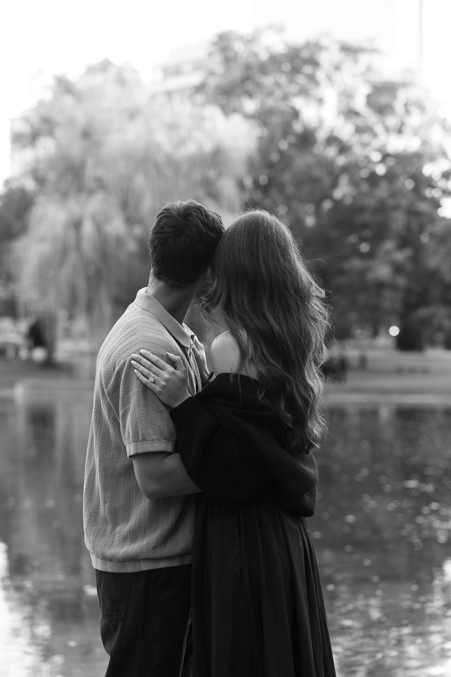 A black and white portrait of a couple embracing by a pond with willow trees in the background.