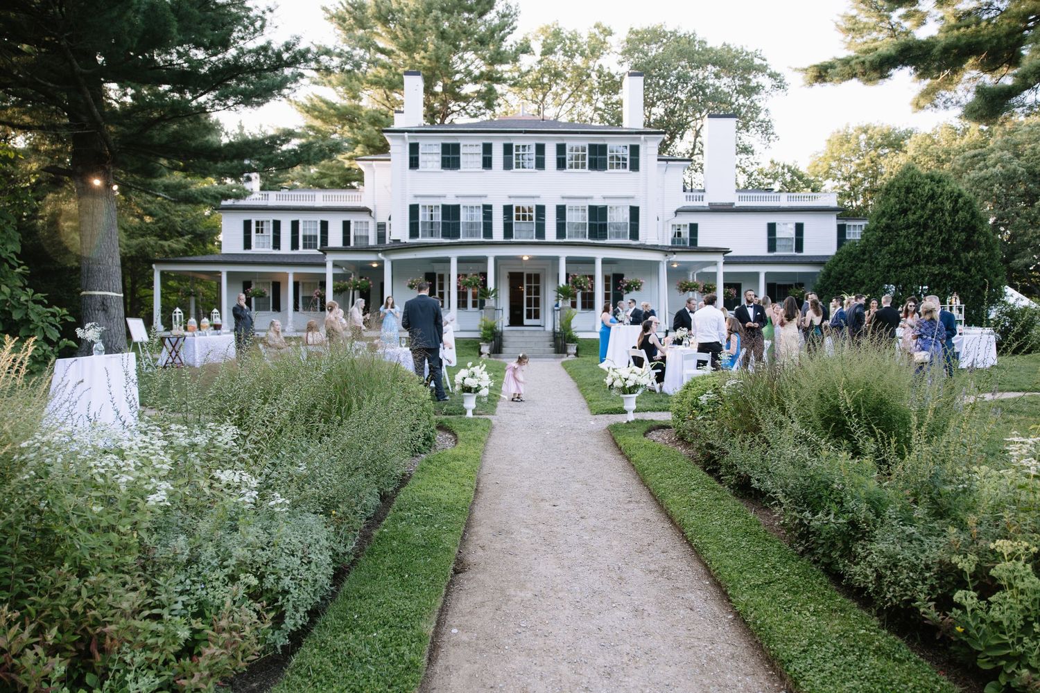 Wedding guests gathering outside a stately white colonial-style mansion.