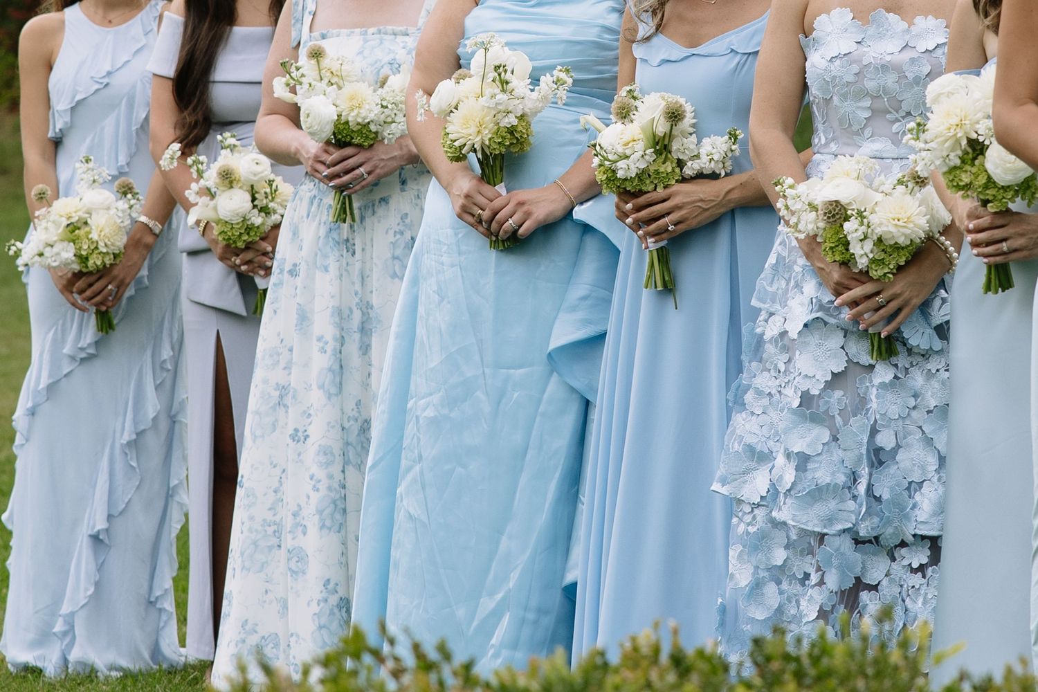 Bridesmaids in light blue dresses hold delicate white and green bouquets during an outdoor wedding.
