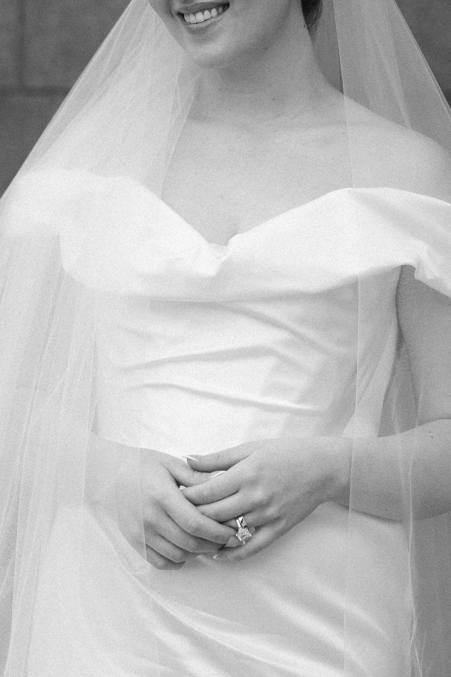 Close-up black and white photo of bride in off-shoulder gown with veil, hands clasped showing diamond ring.