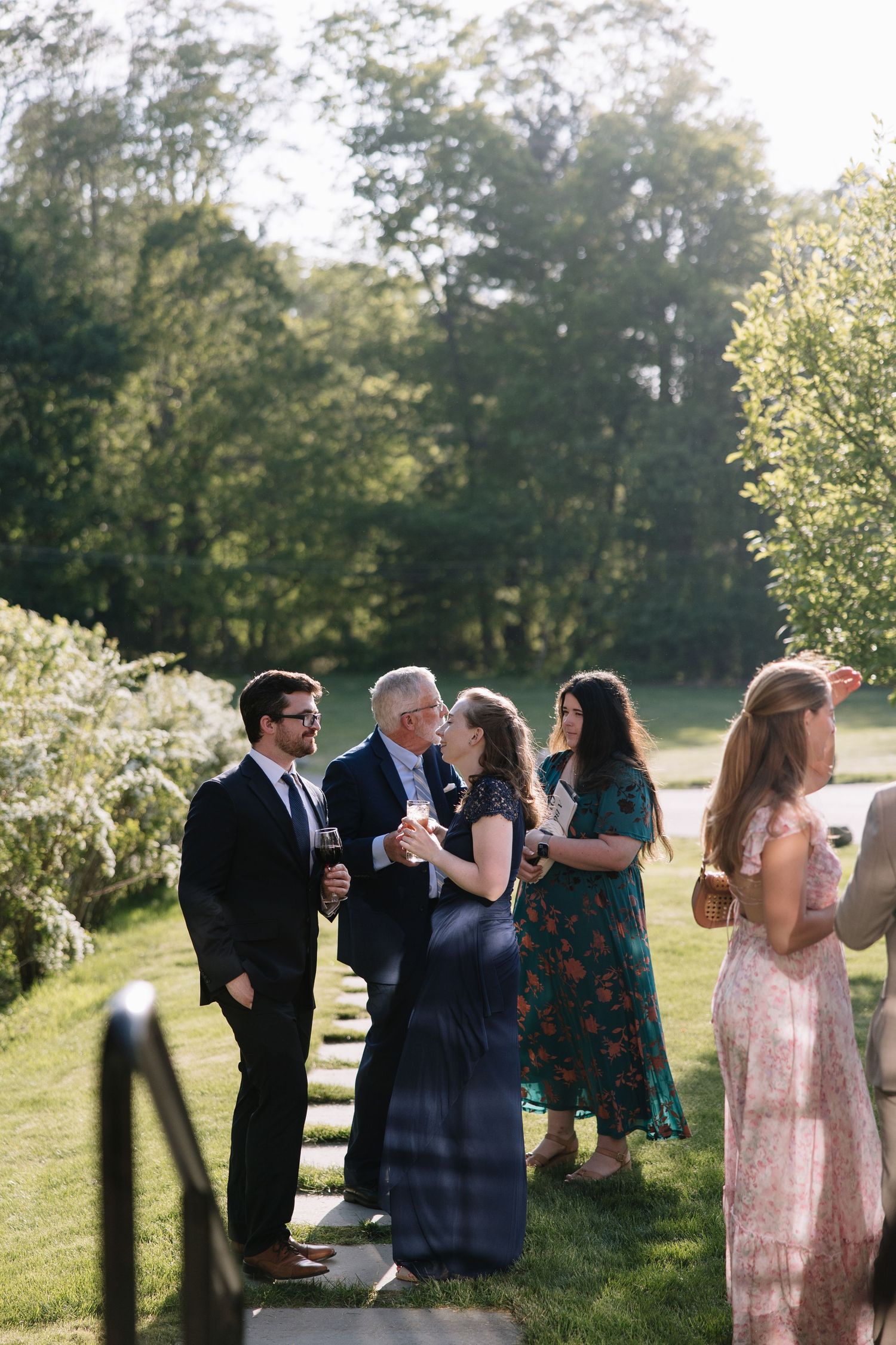 Elegantly dressed wedding guests socialize outdoors on a sunny day amid lush green trees.