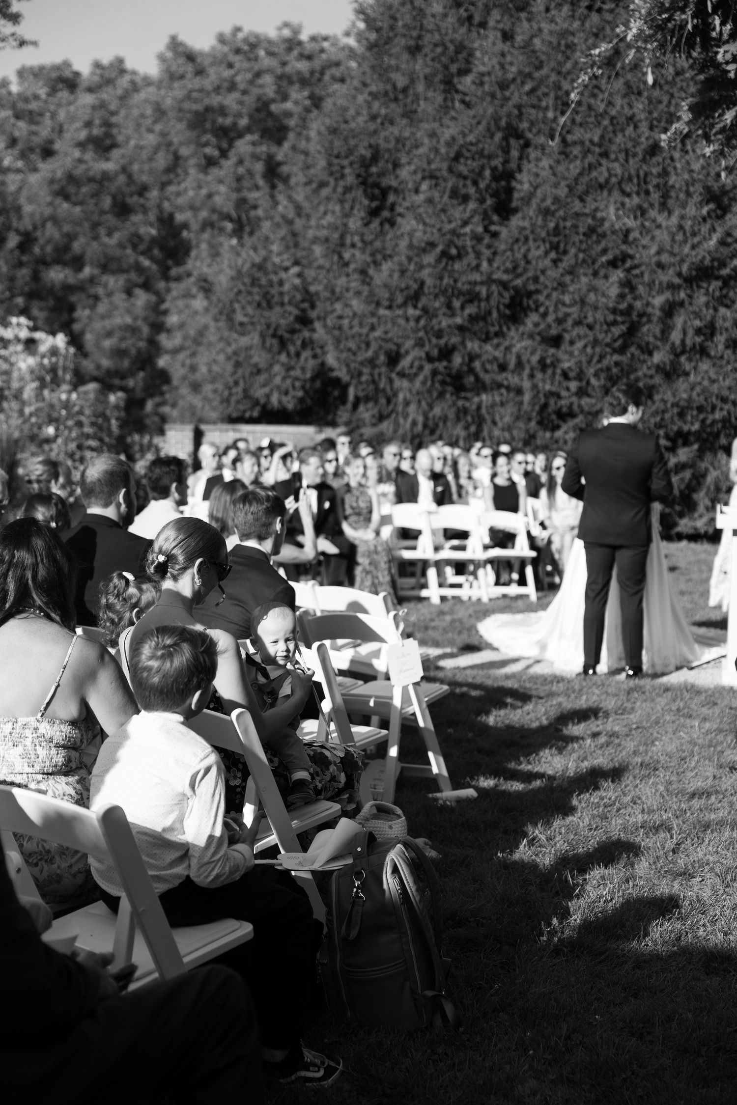 Black and white photo of guests seated in white chairs at an outdoor wedding ceremony on a sunny day.