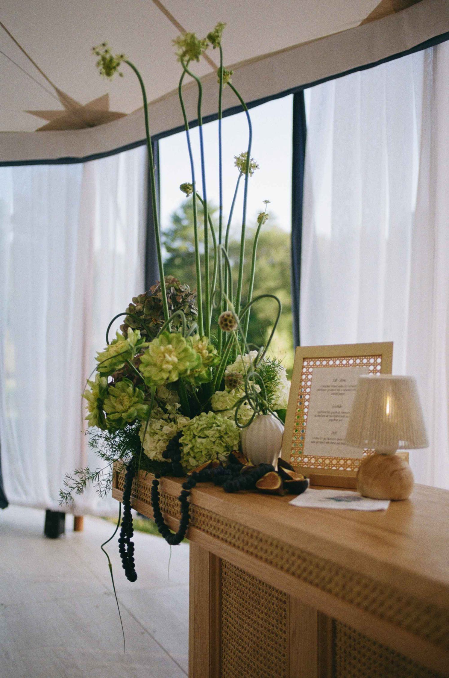 Elegant floral arrangement with green hydrangeas and tall stems on a wooden console table near sheer curtains.