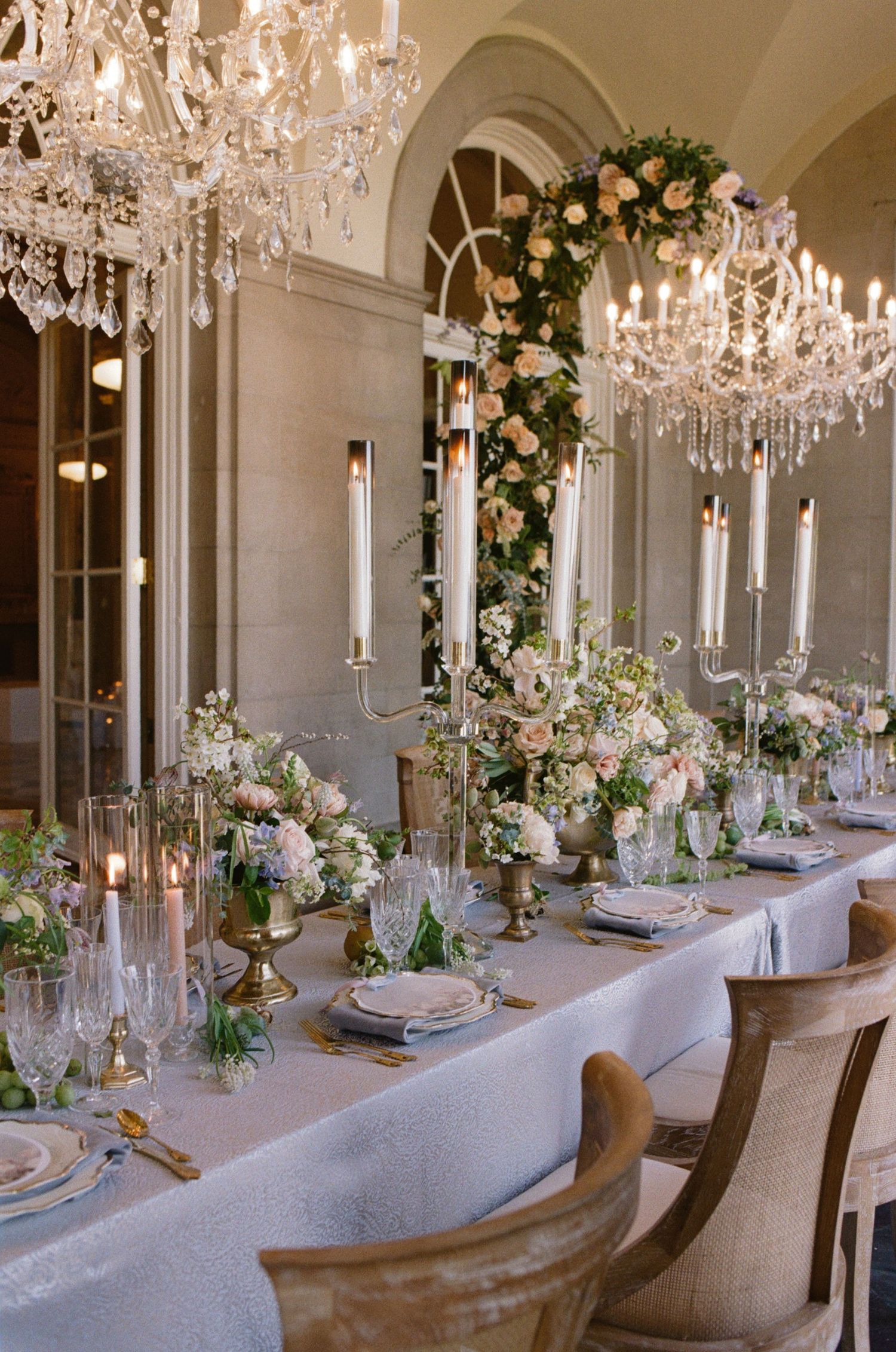 Elegant wedding reception table with crystal chandeliers, floral arrangements, candelabras, and fine china on a white linen tablecloth.