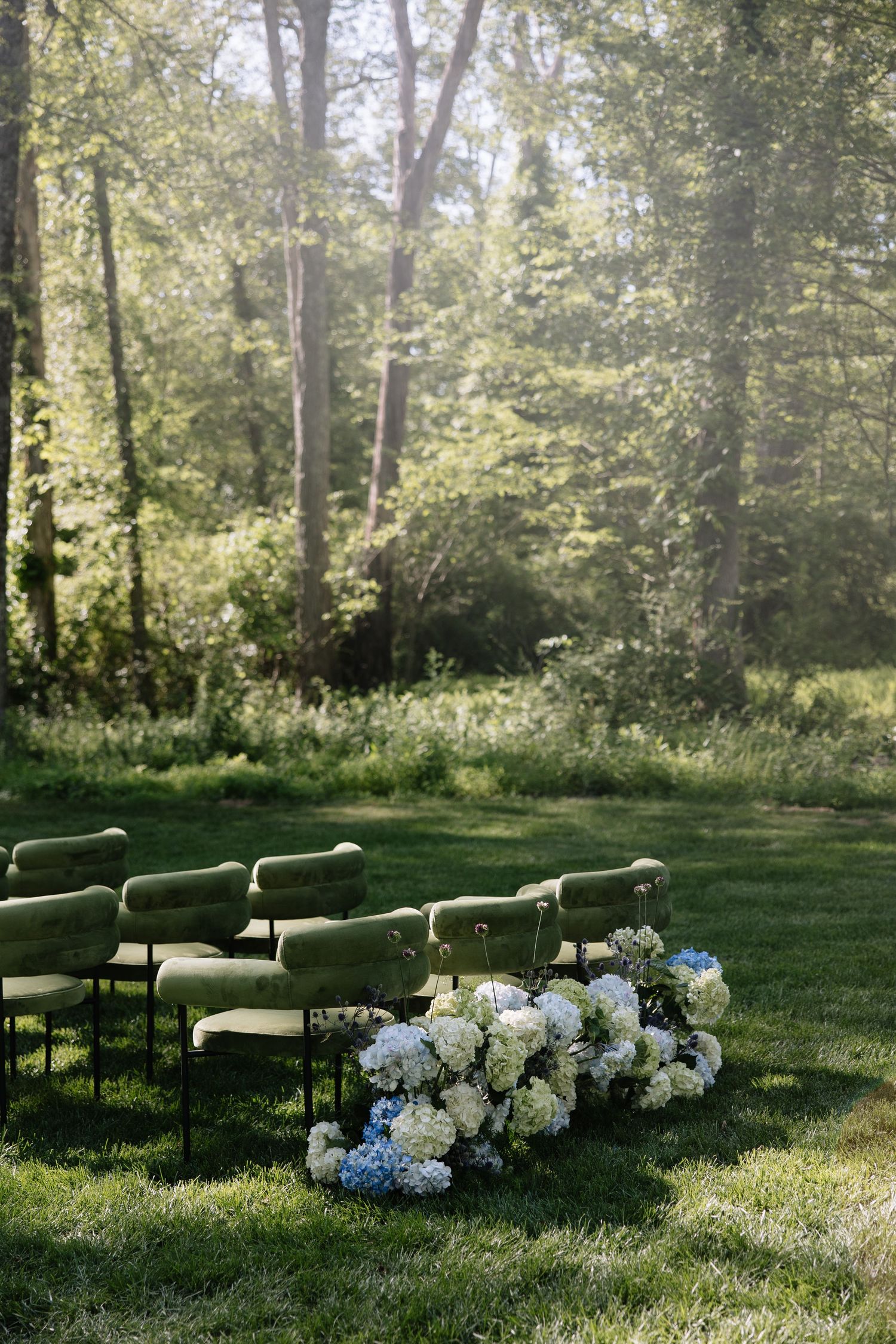Outdoor wedding ceremony setup with green velvet chairs and white and blue hydrangea floral arrangements.