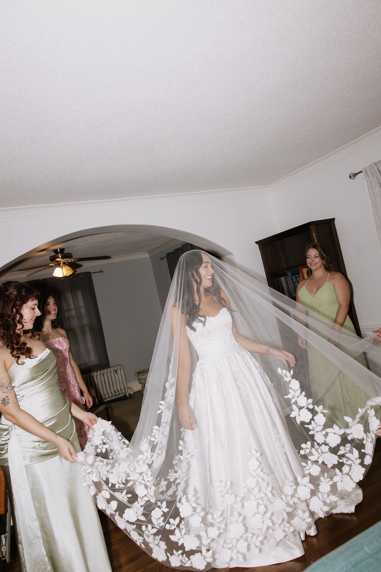 Bride in a white ballgown with floral lace veil held by bridesmaids in sage green dresses inside a home.