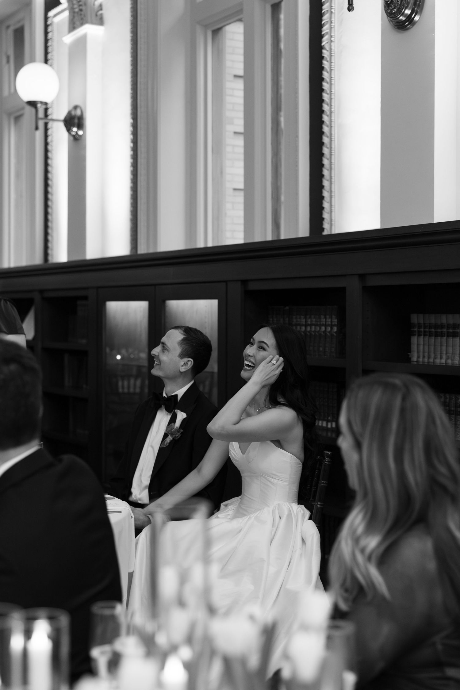 Black and white photo of bride smiling joyfully at wedding reception dinner in elegant library venue.