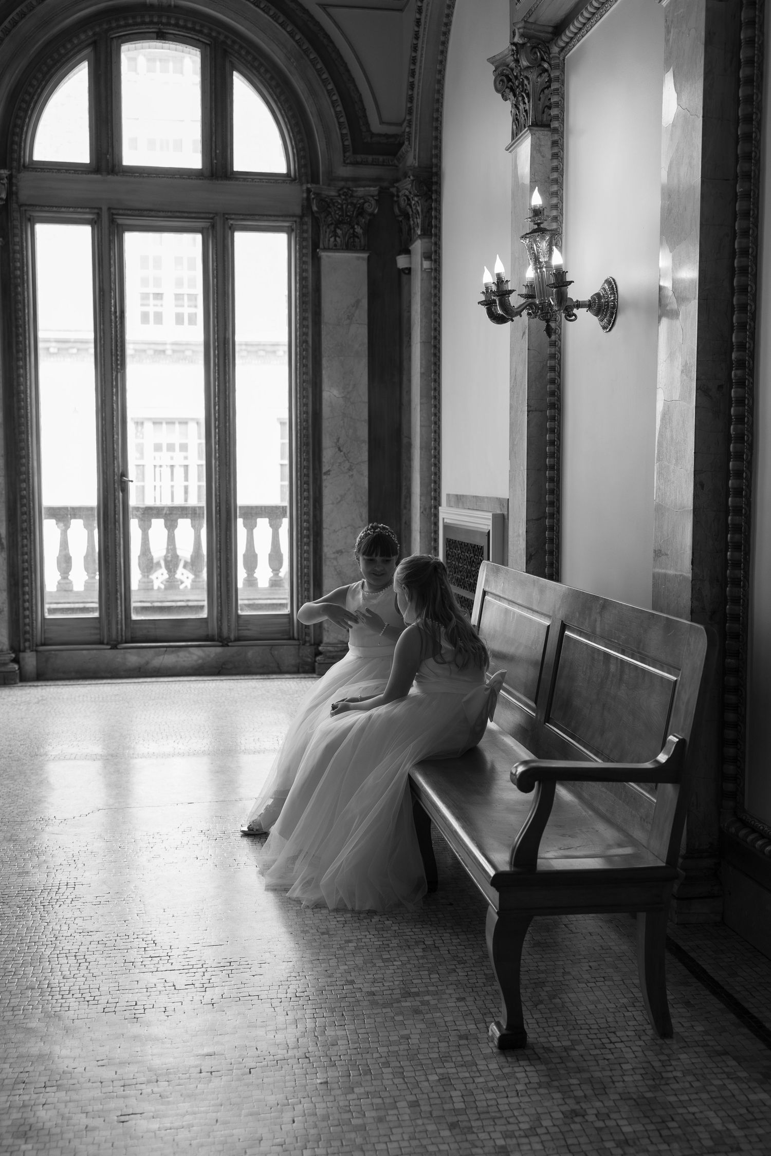 Two brides in white gowns sit together on a wooden bench in an elegant hall with arched windows, black and white photo.