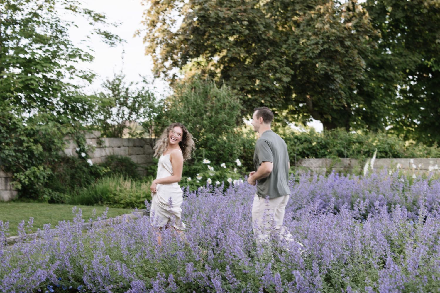 Couple smiling and holding hands in a lush garden surrounded by blooming purple lavender flowers on a sunny day.